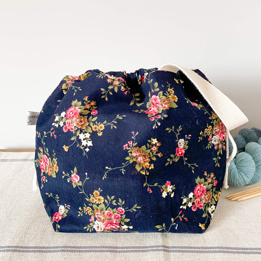 A pretty floral project bag for fibre projects sits on a table next to two skeins of yarn and some wooden knitting needles. 