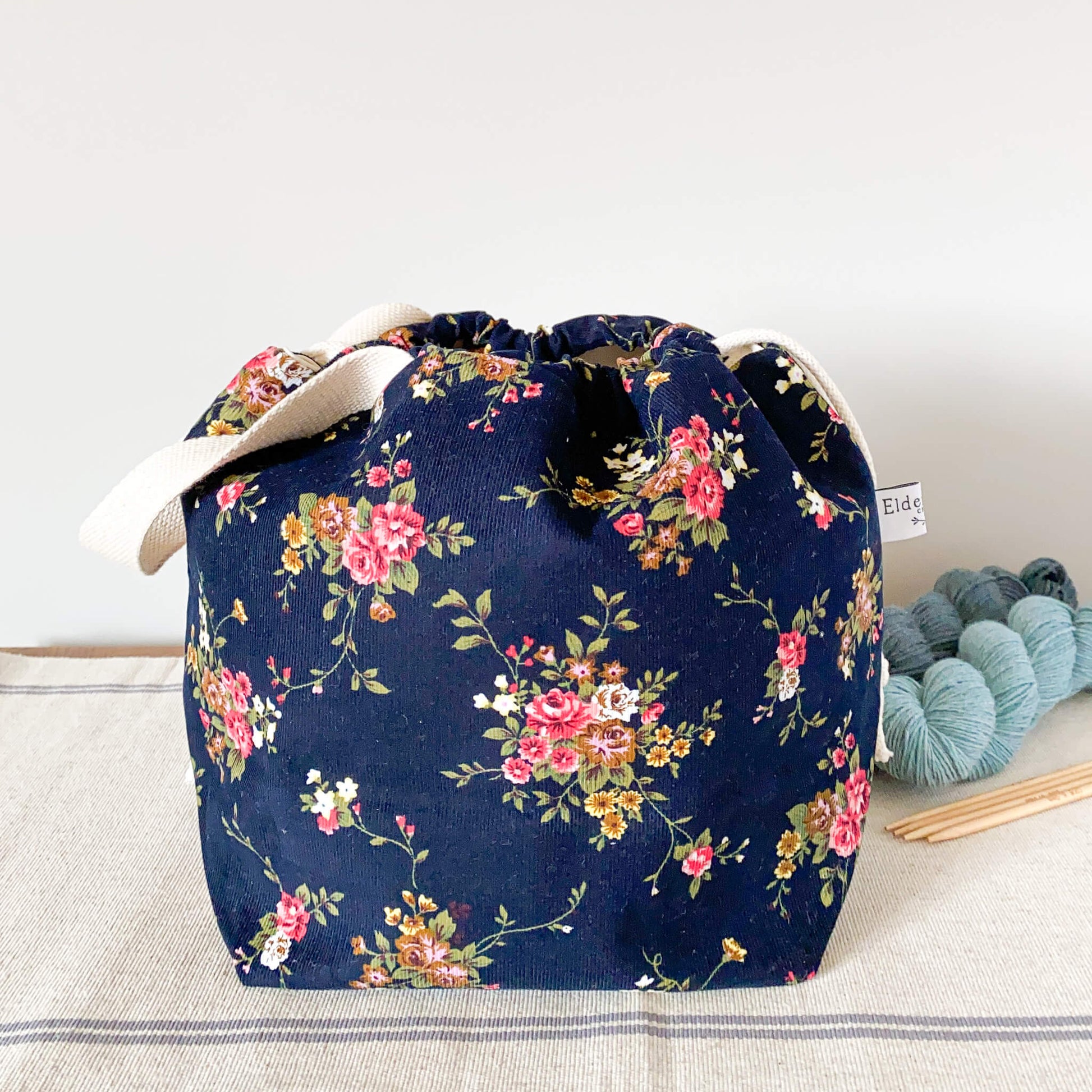 A deep blue project bag sits on a table next to two skeins of yarn. The blue fabric is printed with lots of small leaves and berries and acorns. The top of the bag is pulled closed by a drawstring. 