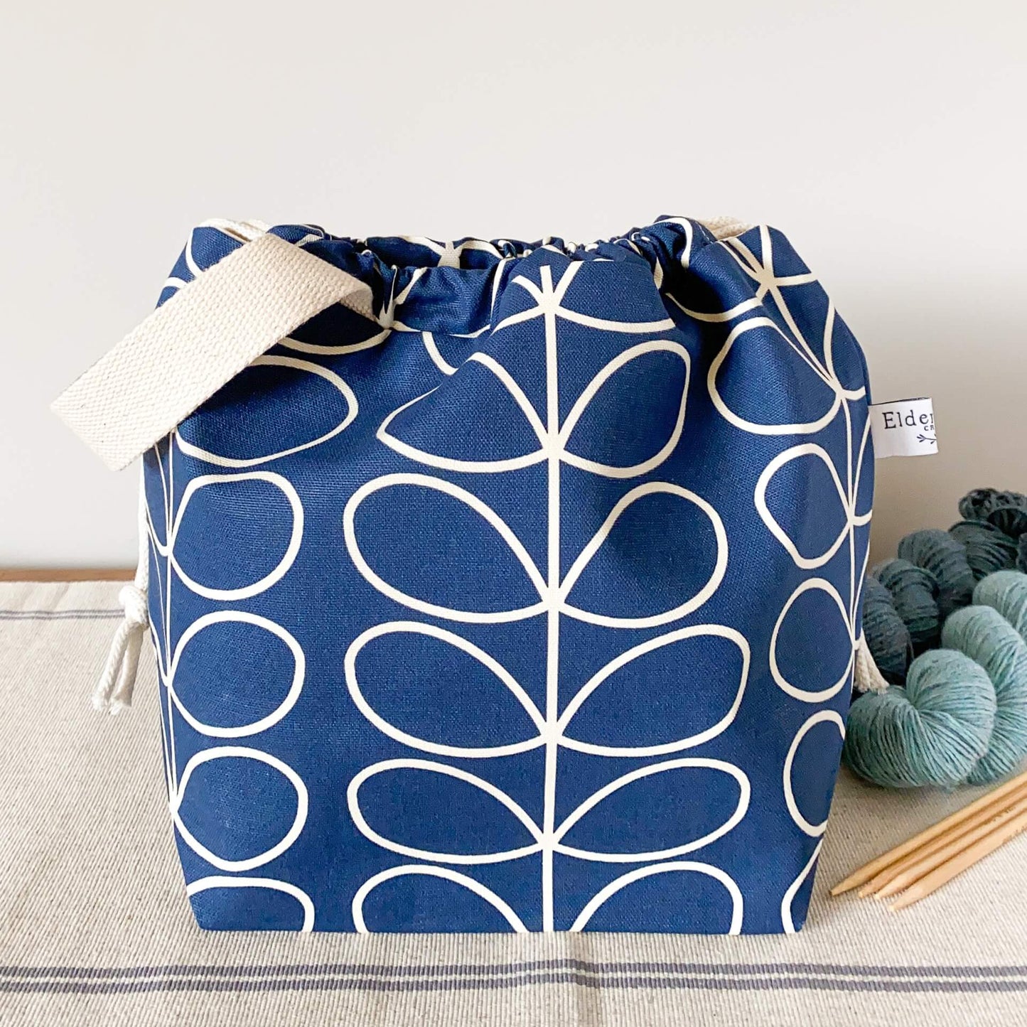 A large blue knitting project bag, pulled closed hiding its contents, sits in front of some yarn and knitting needles.