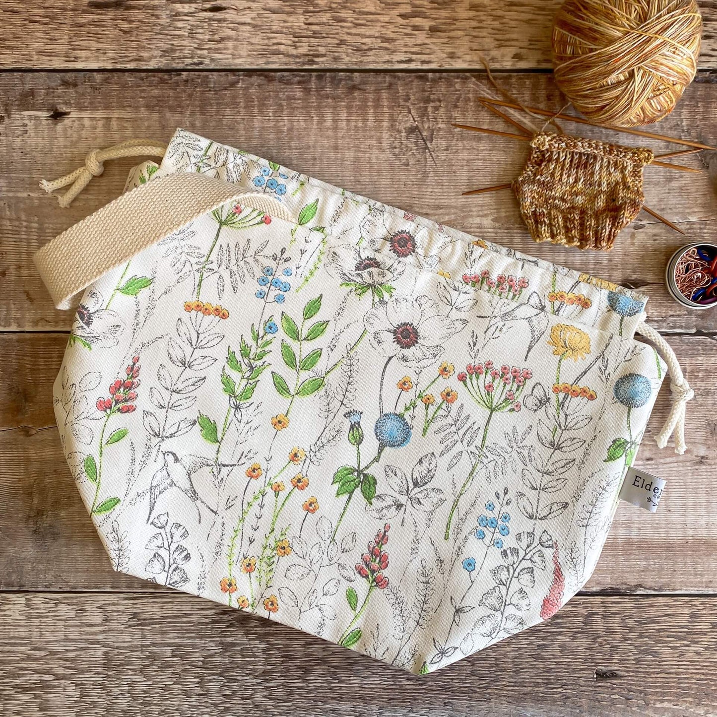 A summery print knitting project bag lies on a wooden table next to an in progress knitting project and a tin of stitch markers.