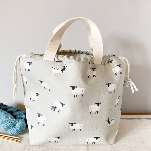A large drawstring bag designed to hold knitting projects sits on a table next to two skeins of yarn and some wooden knitting needles. The bag features a sheepy pring fabric. 
