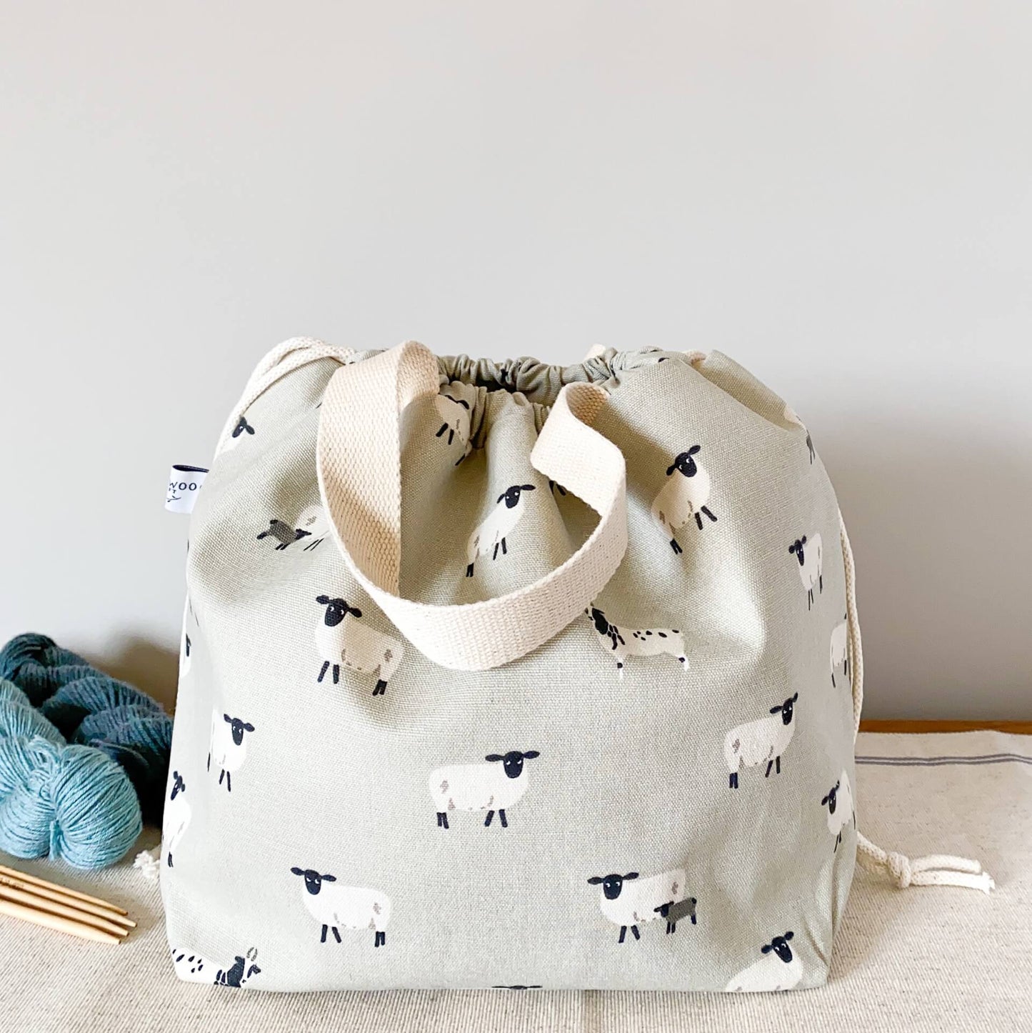 A large drawstring bag designed to hold knitting projects sits on a table next to two skeins of yarn and some wooden knitting needles. The bag features a sheepy pring fabric. The bag is pulled closed by a drawstring hiding its contents.
