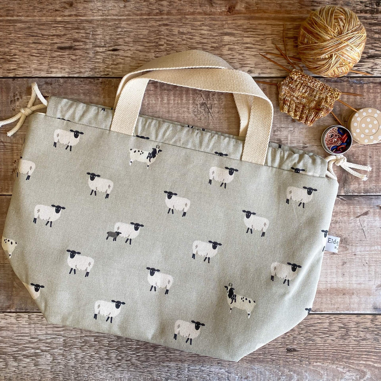 A sheepy project bag for knitting lies on a wooden table next to a knitting project - possibly a sock - and a tin of stitch markers.