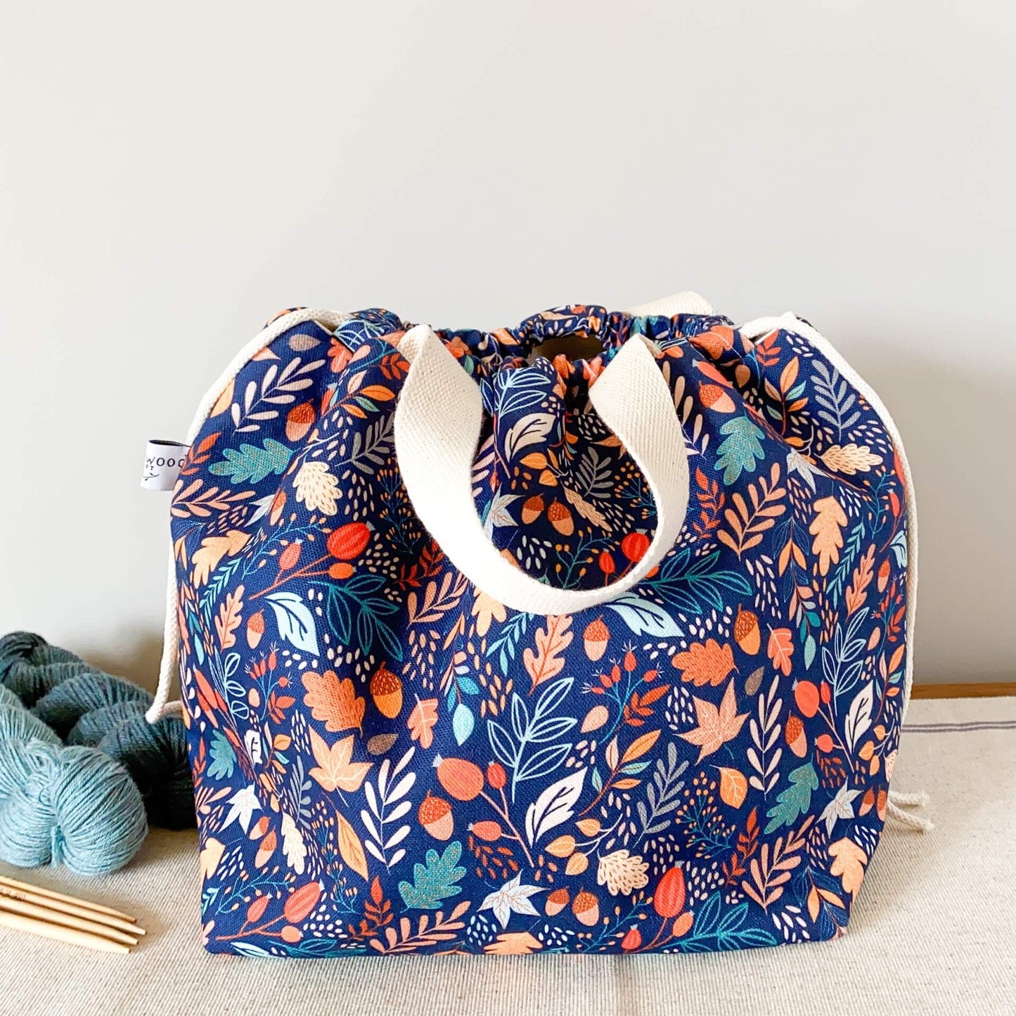 A knitting project bag sits on a table. The bag is made from a fabric that features a bright leaf and acorn print. The top of the bag is pulled closed hiding the contents of the bag. 