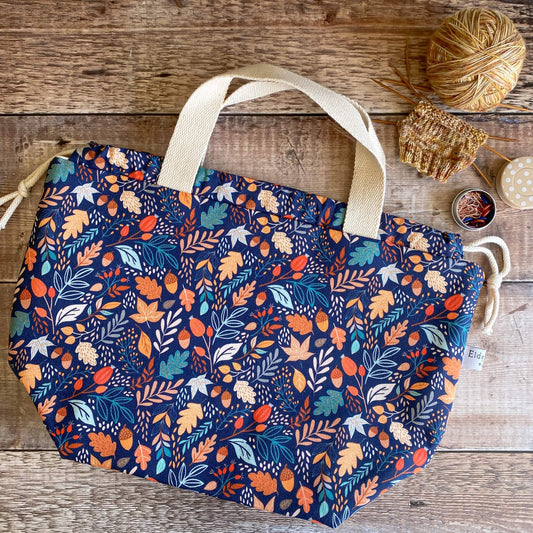 A large knitting project bag featuring a bright leaf print on a blue background, lies on a wooden table next to an in progress knitting project and a tin of stitch markers. 