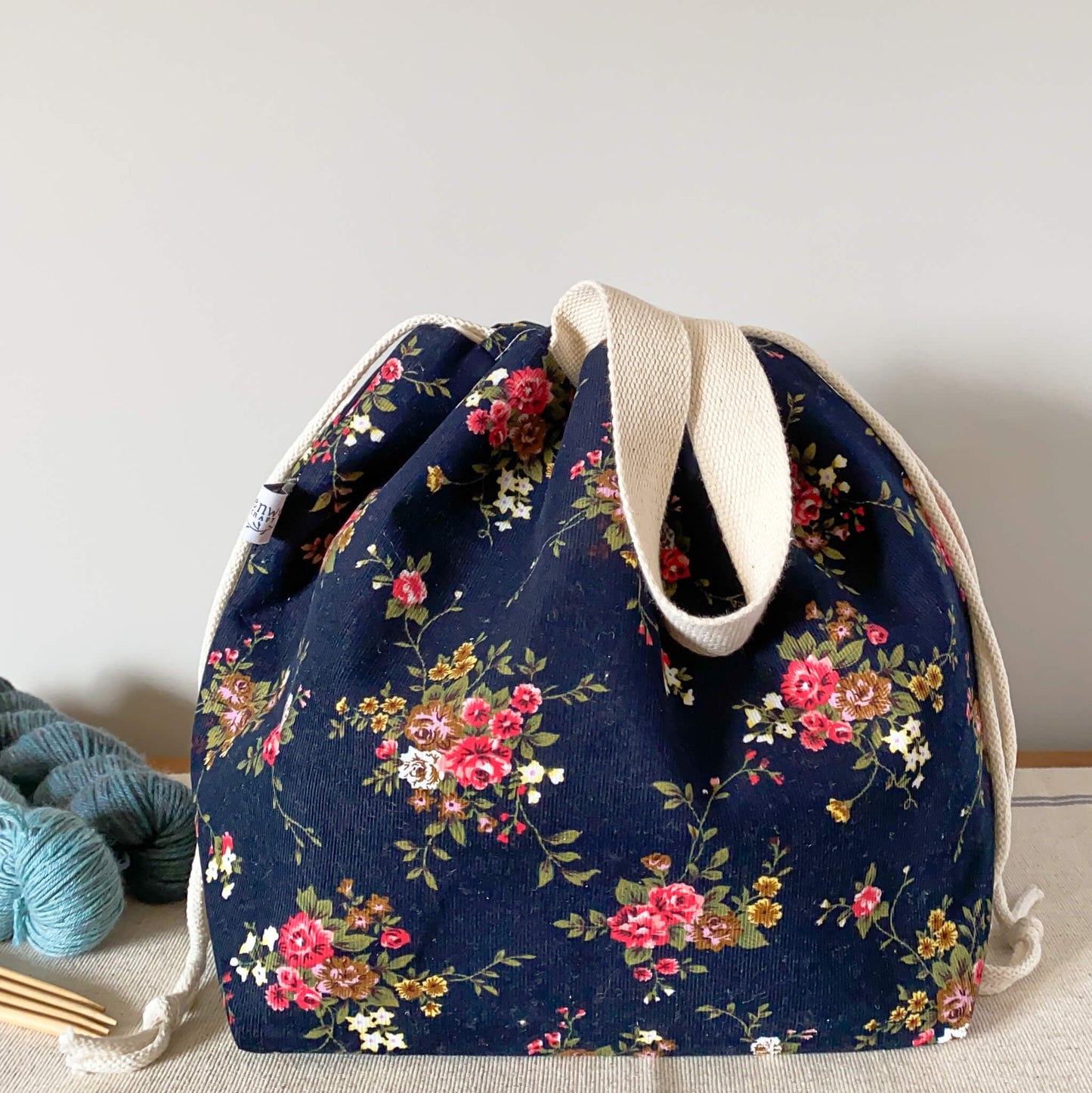 A pretty floral knitting project bag sits on a table next to two skeins of yarn and some wooden knitting needles. The bag is pulled closed hiding the contents. 