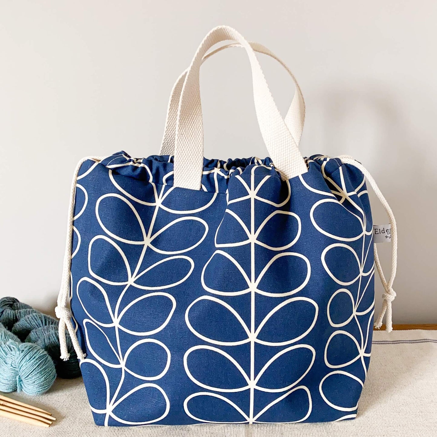 A large blue knitting project bag sits on a table next to a small pile of yarn and some wooden knitting needles.