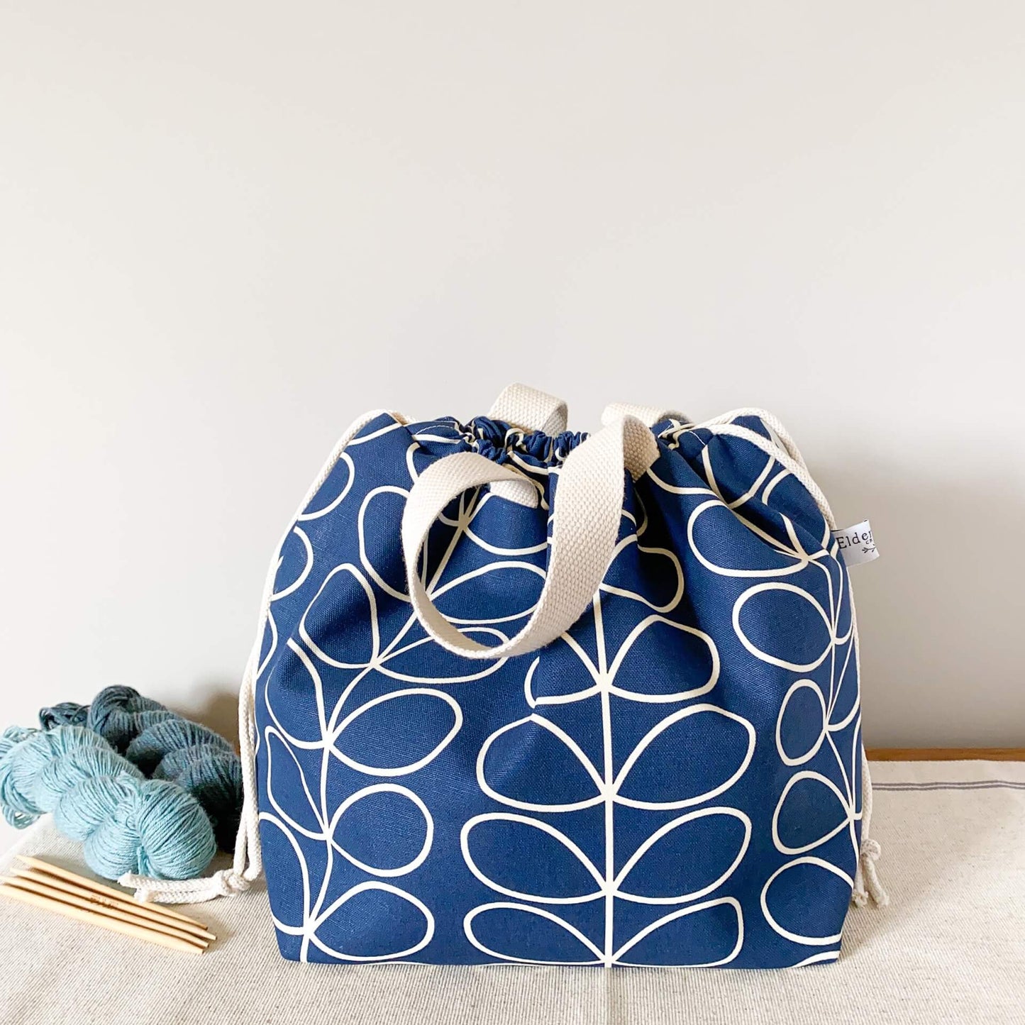 A large blue knitting proect bag sits on a table next to some yarn and knitting needles. The bag is pulled closed hiding the contents.