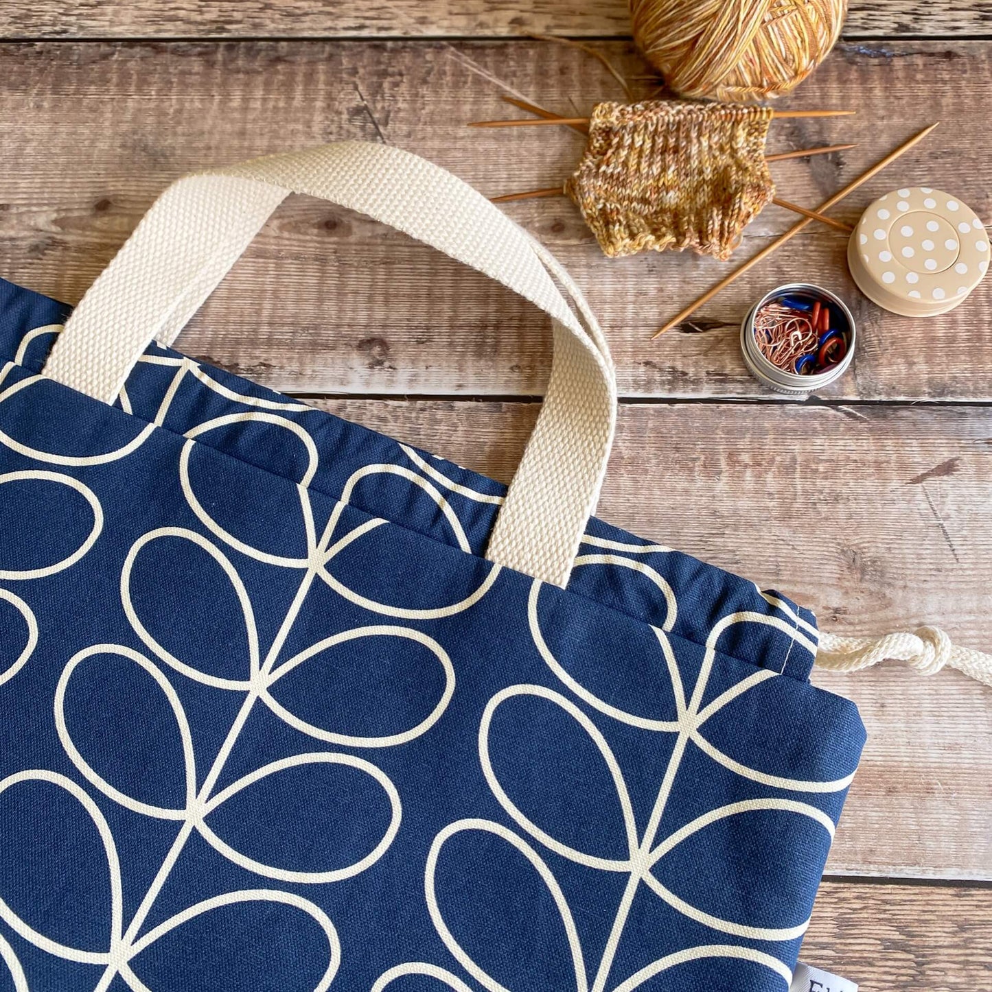 A blue and white project bag lies flat on a wooden table. Next to the bag is a small knitting project and a tin of stitch markers.