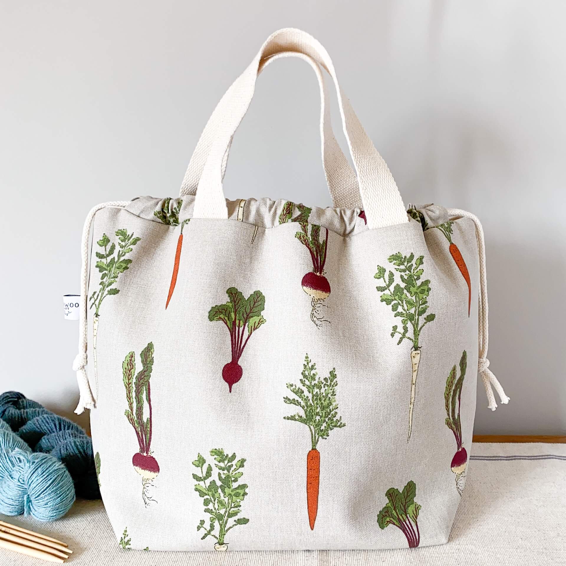 A large knitting project bag featuring a home grown vegetable print fabric, sits on a table next to two skeins of yarn and a set of wooden knitting needles. 