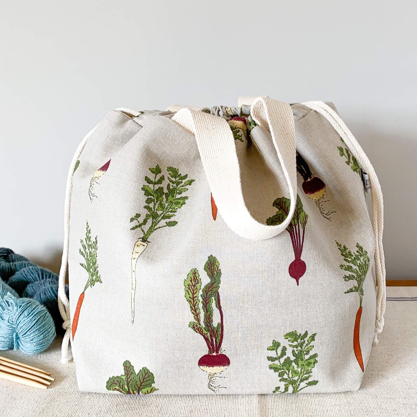 A large knitting project bag sits on a table next to some blue yarn and wooden knitting needles. The top of the bag is pulled closed hiding the contents and the bag's handles hang down in front of the bag.