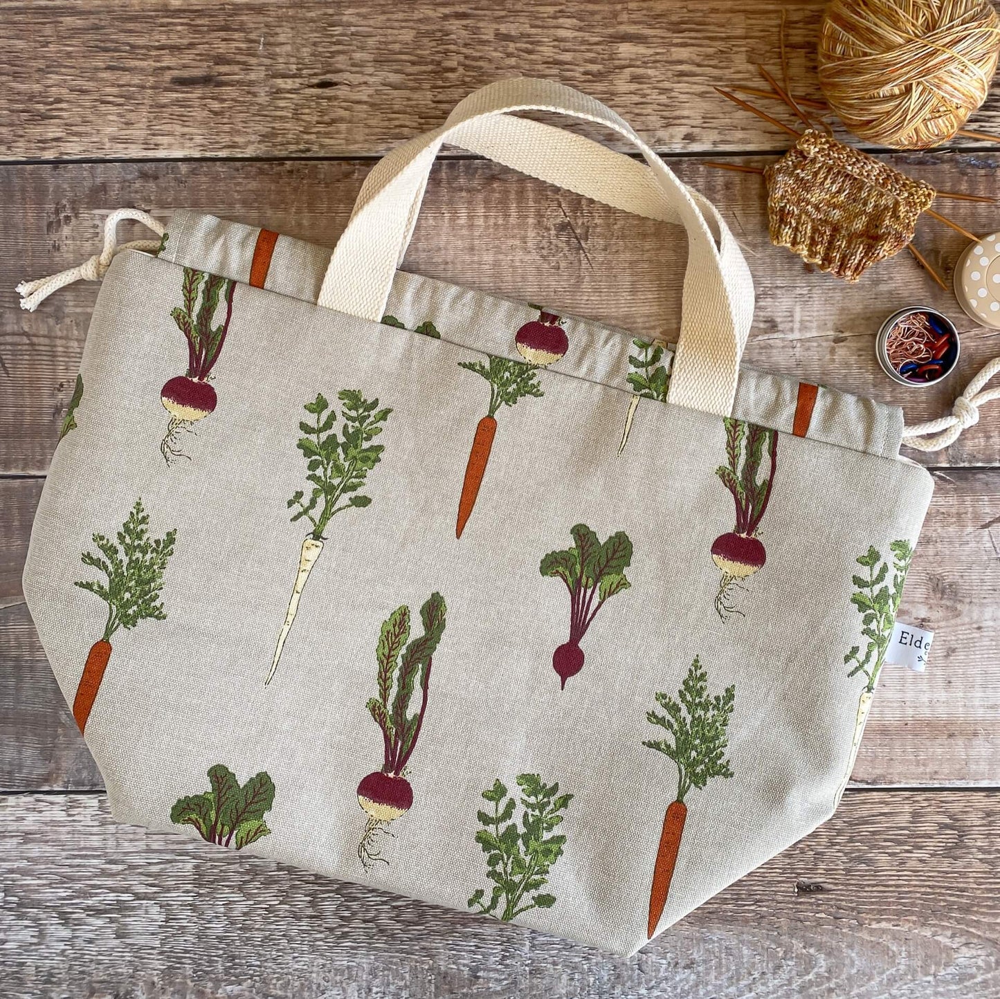 A knitting project bag made from a vegetable print fabric lies flat on a table next to a small knitting project.