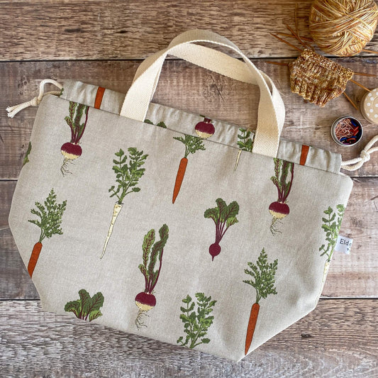 A knitting project bag made from a vegetable print fabric lies flat on a table next to a small knitting project. 