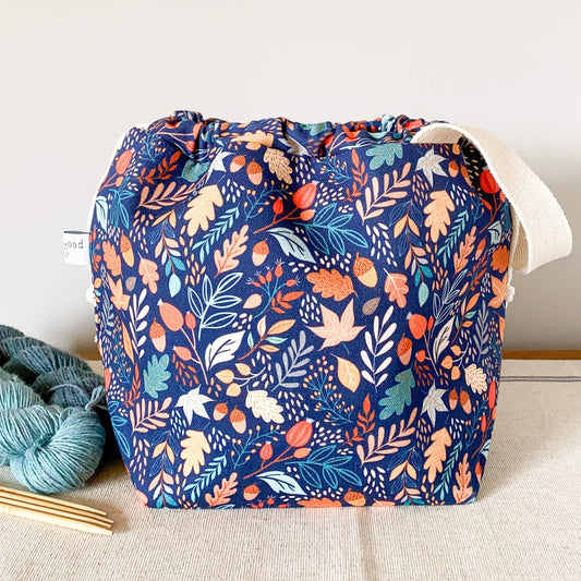 A deep blue project bag sits on a table next to two skeins of yarn. The blue fabric is printed with lots of small leaves and berries and acorns. 