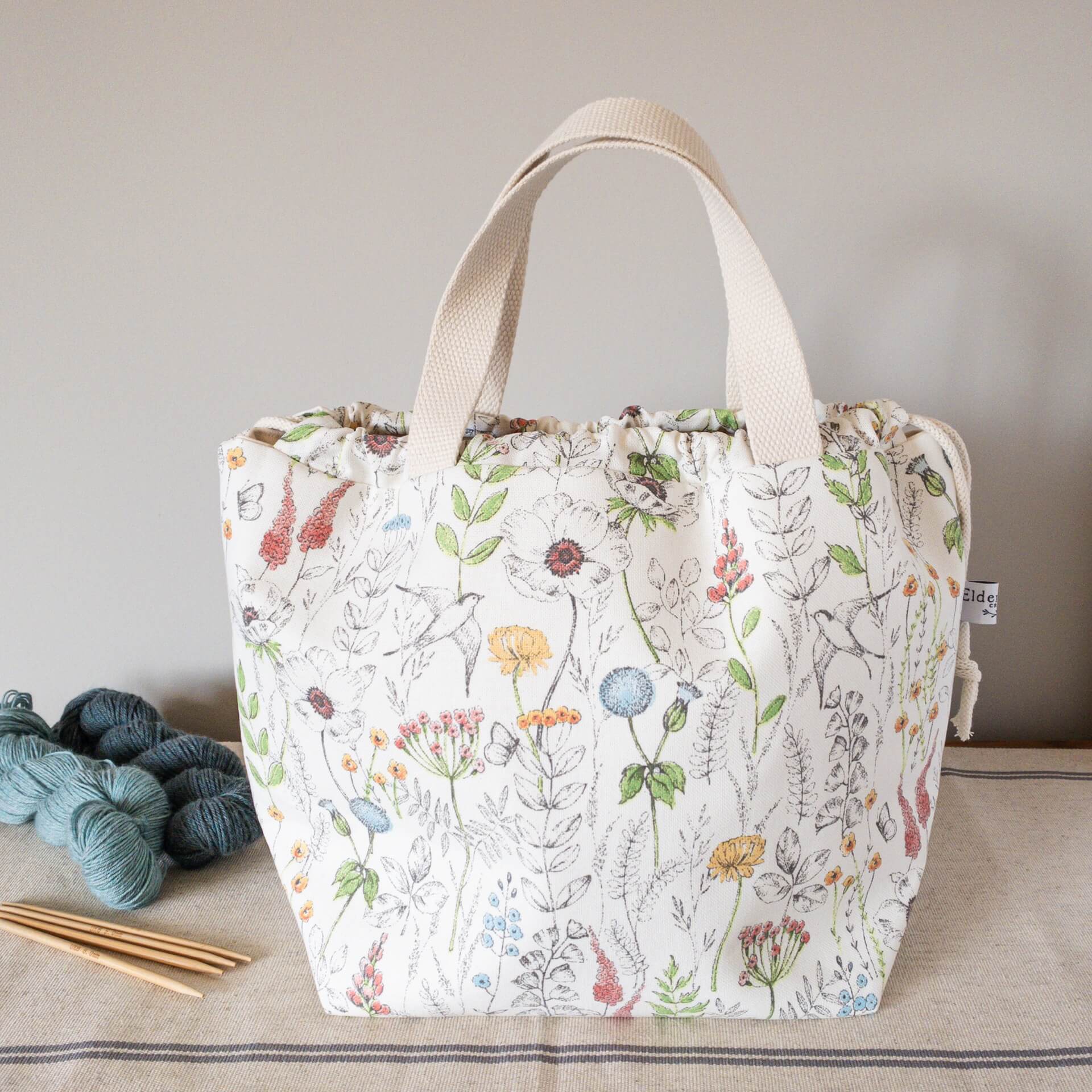 A large knitting project bag sits on a wooden table next to two skeins of yar and some wooden knitting needles. The bag is made from a summery floral fabric. 