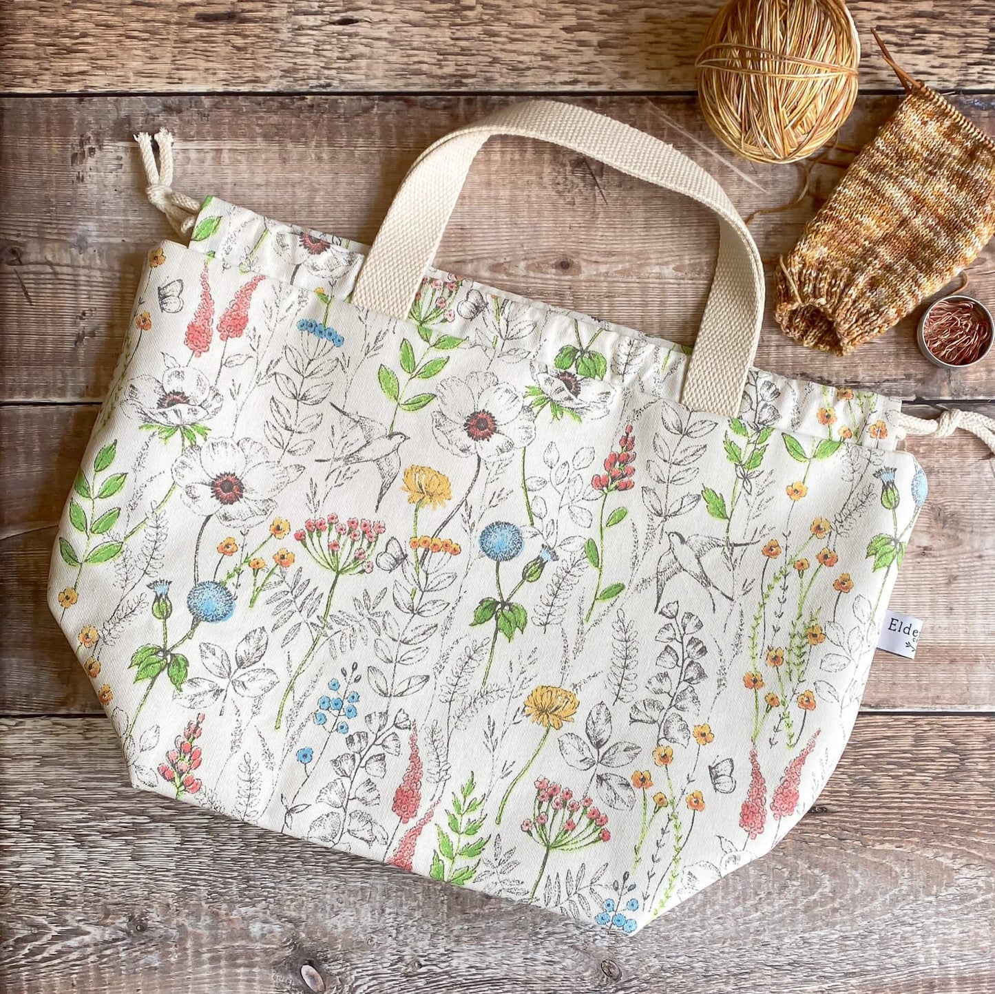 A large knitting project bag in a summery floral print sits on a wooden table next to an in progress knitting project.
