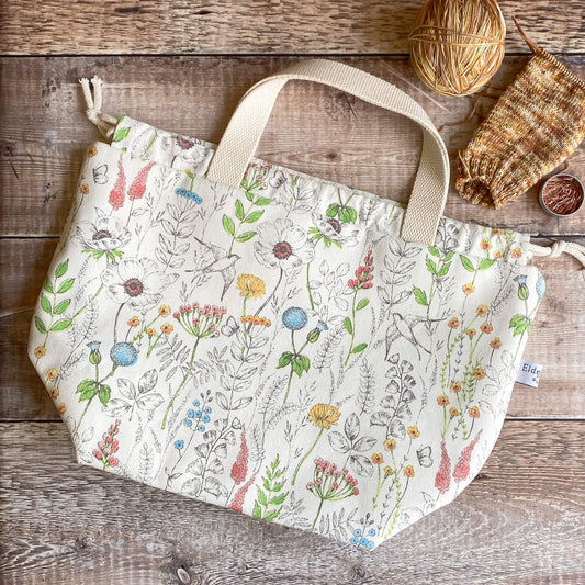 A large knitting project bag in a summery floral print sits on a wooden table next to an in progress knitting project. 