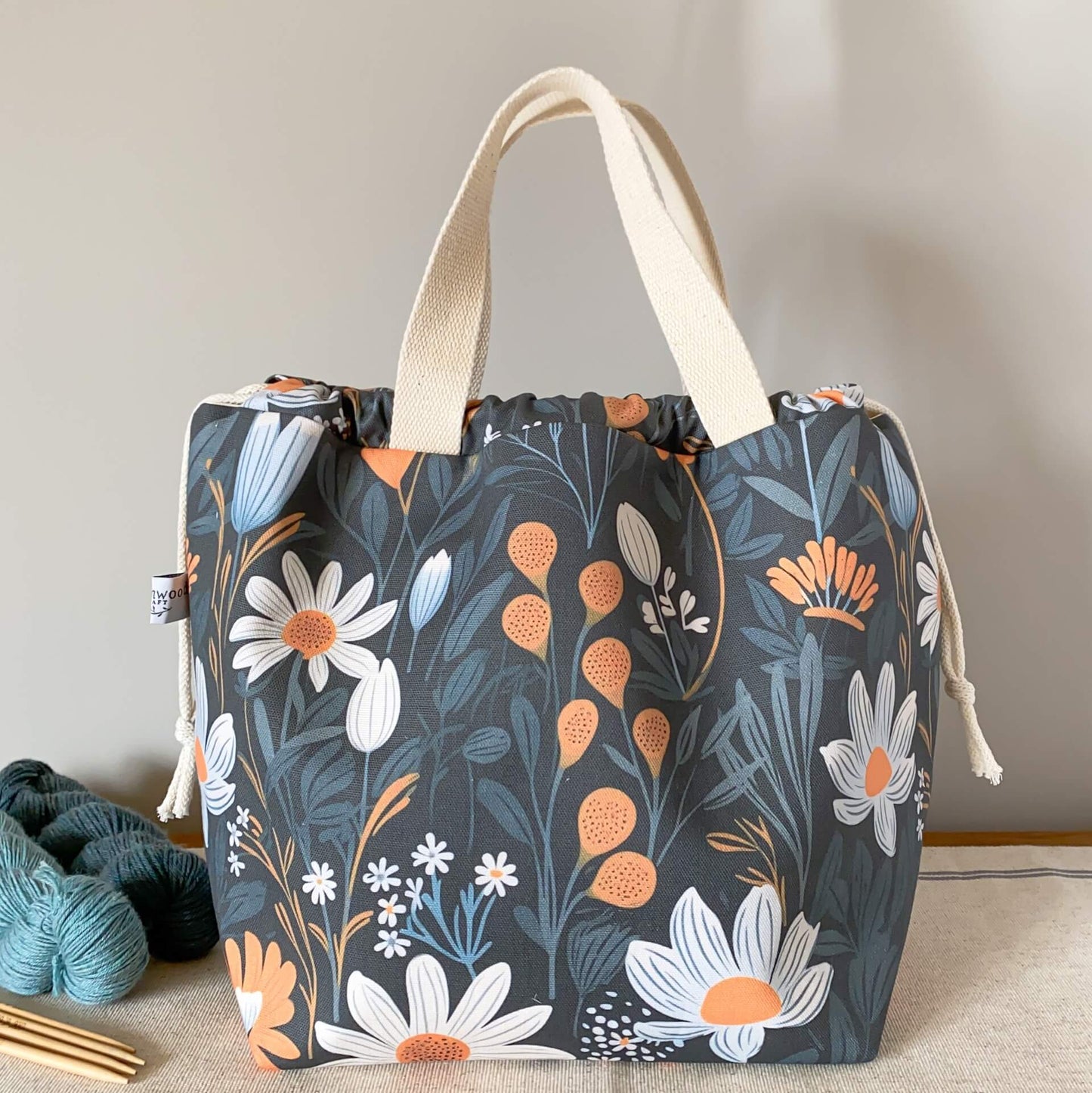 A large knitting project bag featuring a dark and bold fabric, sits on a table next to two skeins of yarn and some wooden knitting needles.