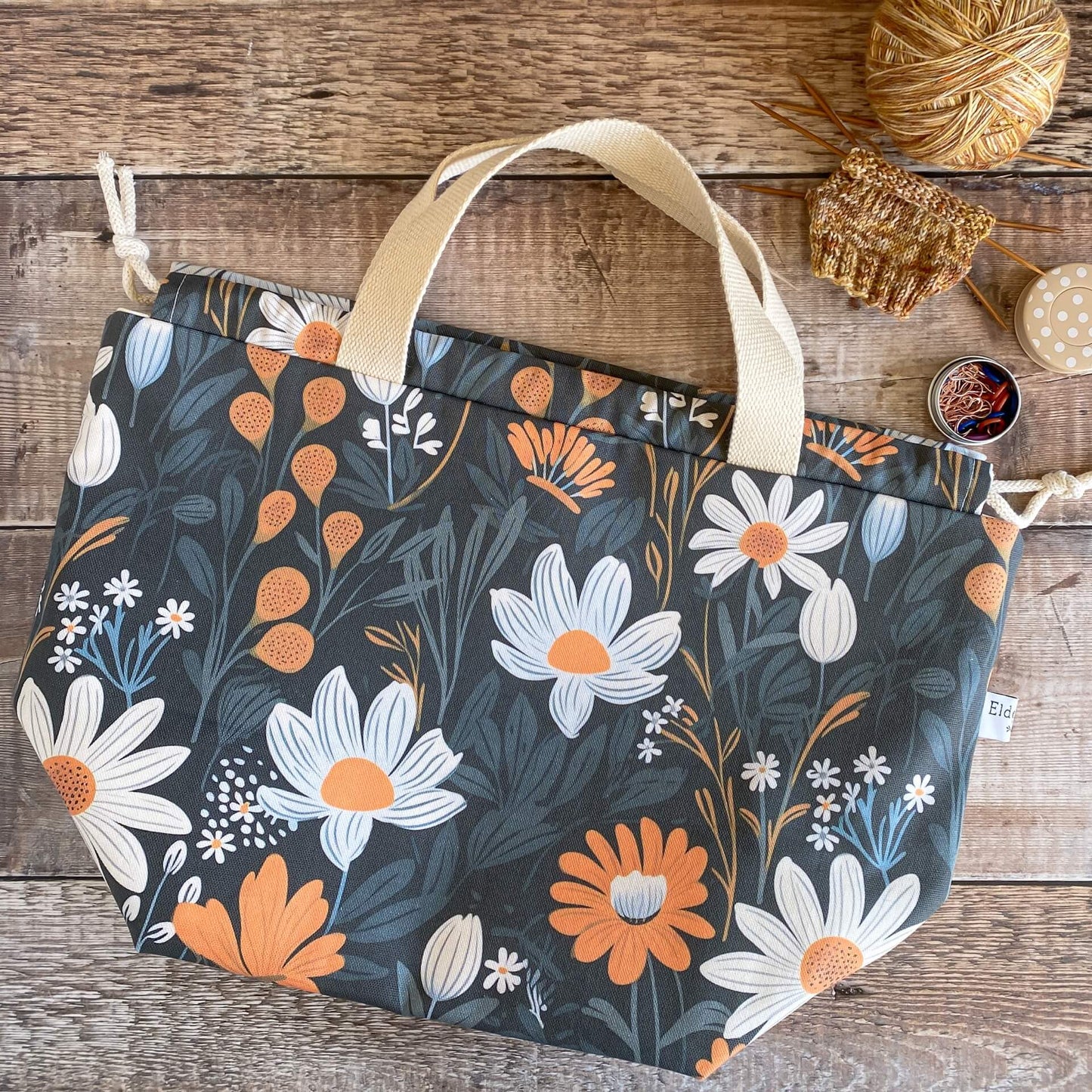 A large knitting project bag lies on a wooden table next to a knitting project. The bag features a dark and bold floral print.