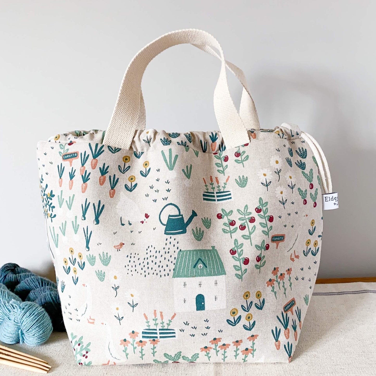 A large knitting project bag sits on a table next to some yarn. The bag is made from cottage garden themed fabric. 