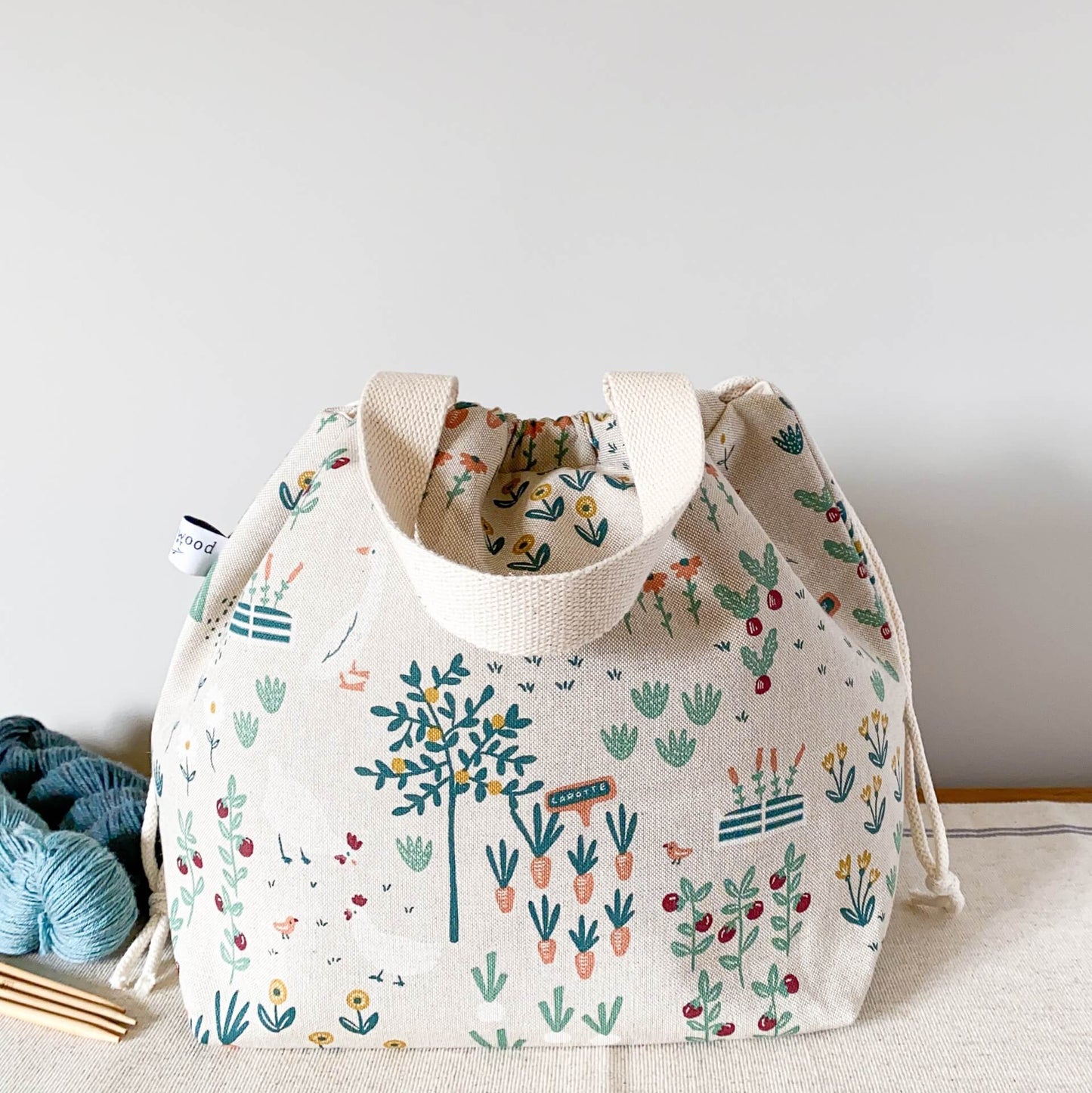 A cottage garden themed fabric sits on a table next to some yarn and knitting needles. The bag is pulled closed hiding its contents.