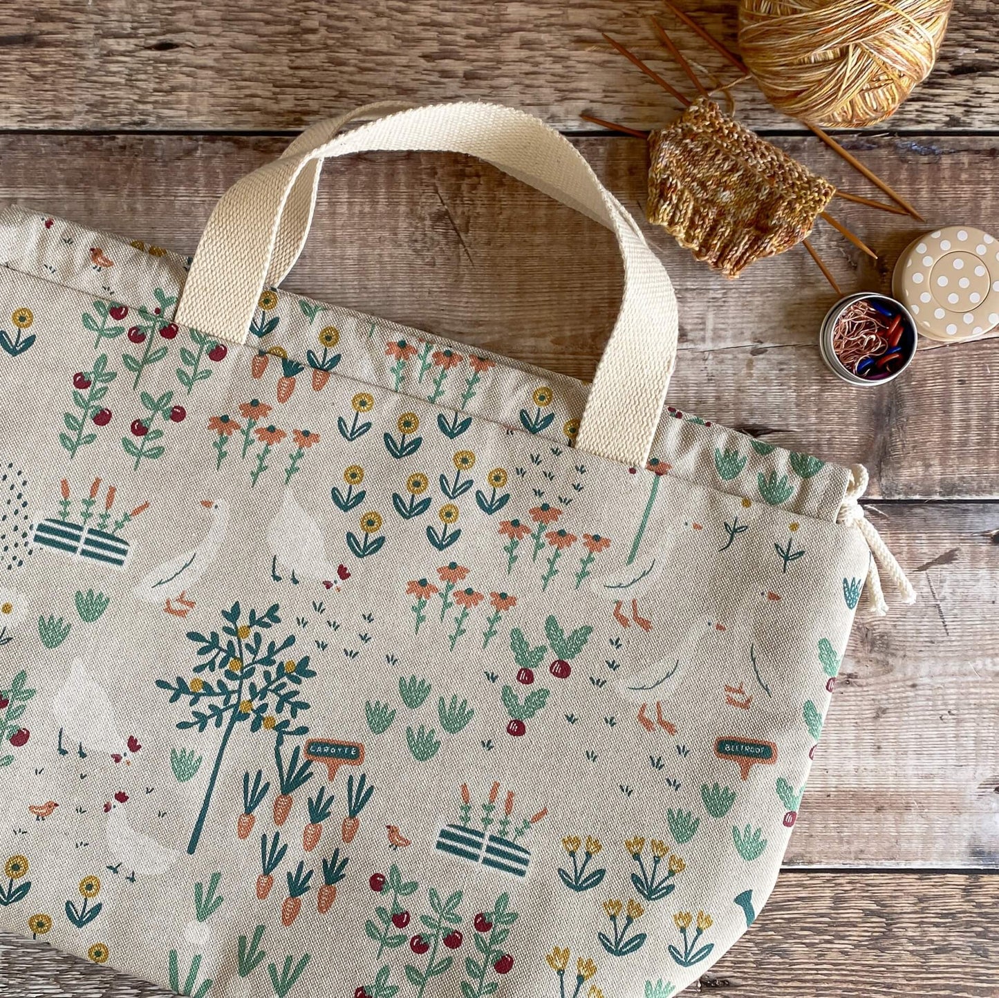 A cottage garden themed project bag lies flat on a wooden table. Next to the bag is a knitting project and a tin of stitch markers and a spotty tape measure.