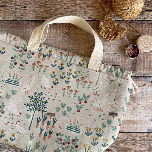A cottage garden themed project bag lies flat on a wooden table. Next to the bag is a knitting project and a tin of stitch markers and a spotty tape measure.