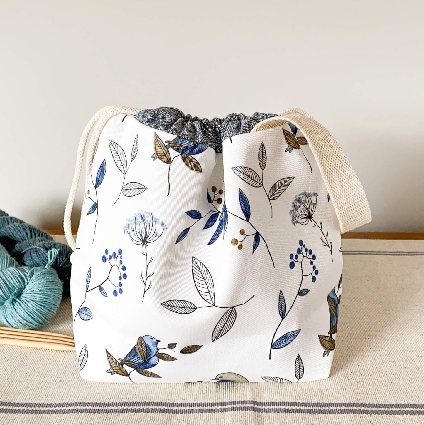 A project bag for knitting sits on a table next to two skeins of blue yarn and a set of wooden needles.