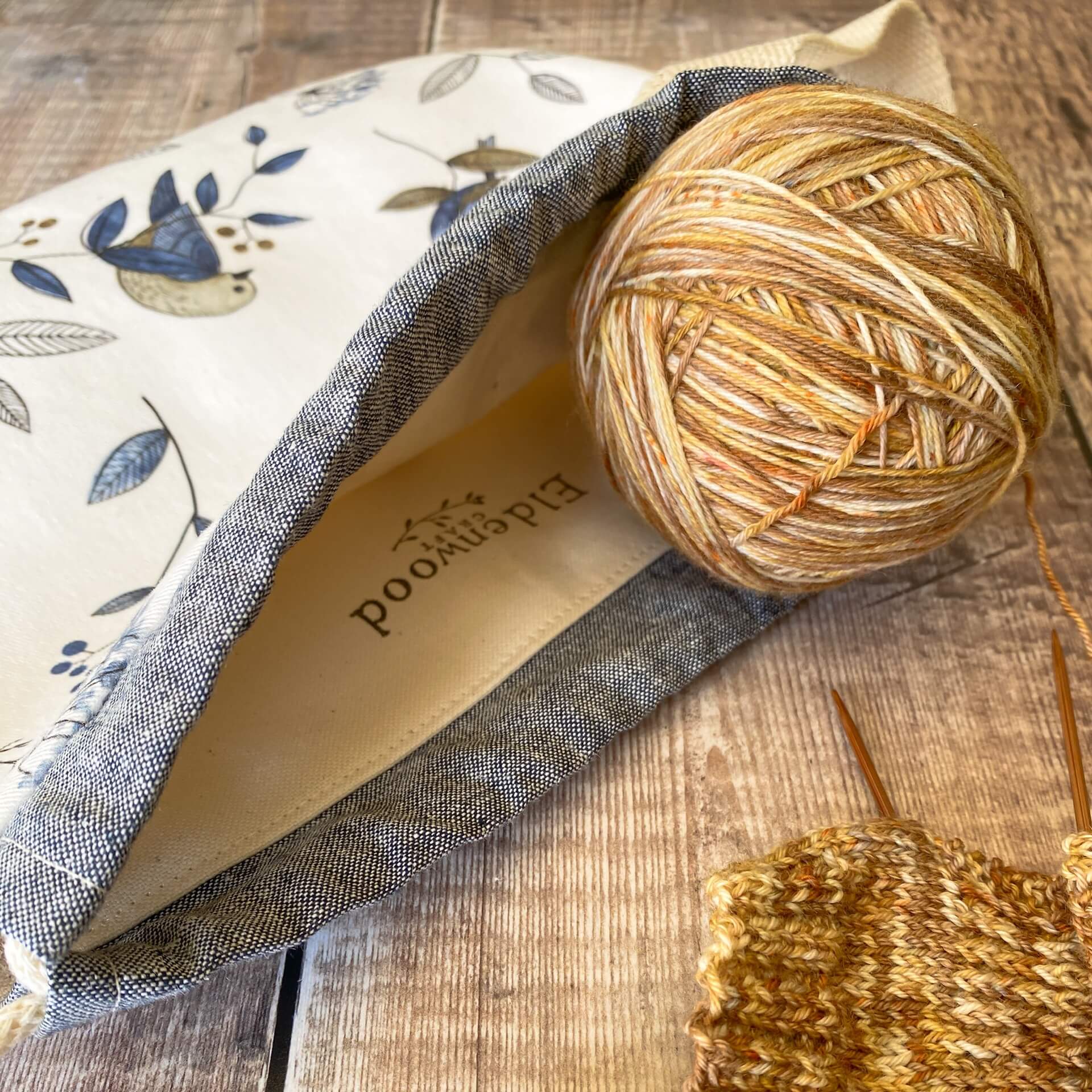 A knitting project bag lies on a table. There is a ball of wool sitting at the opening and we can just see inside to a printed logo that says Eldenwood Craft