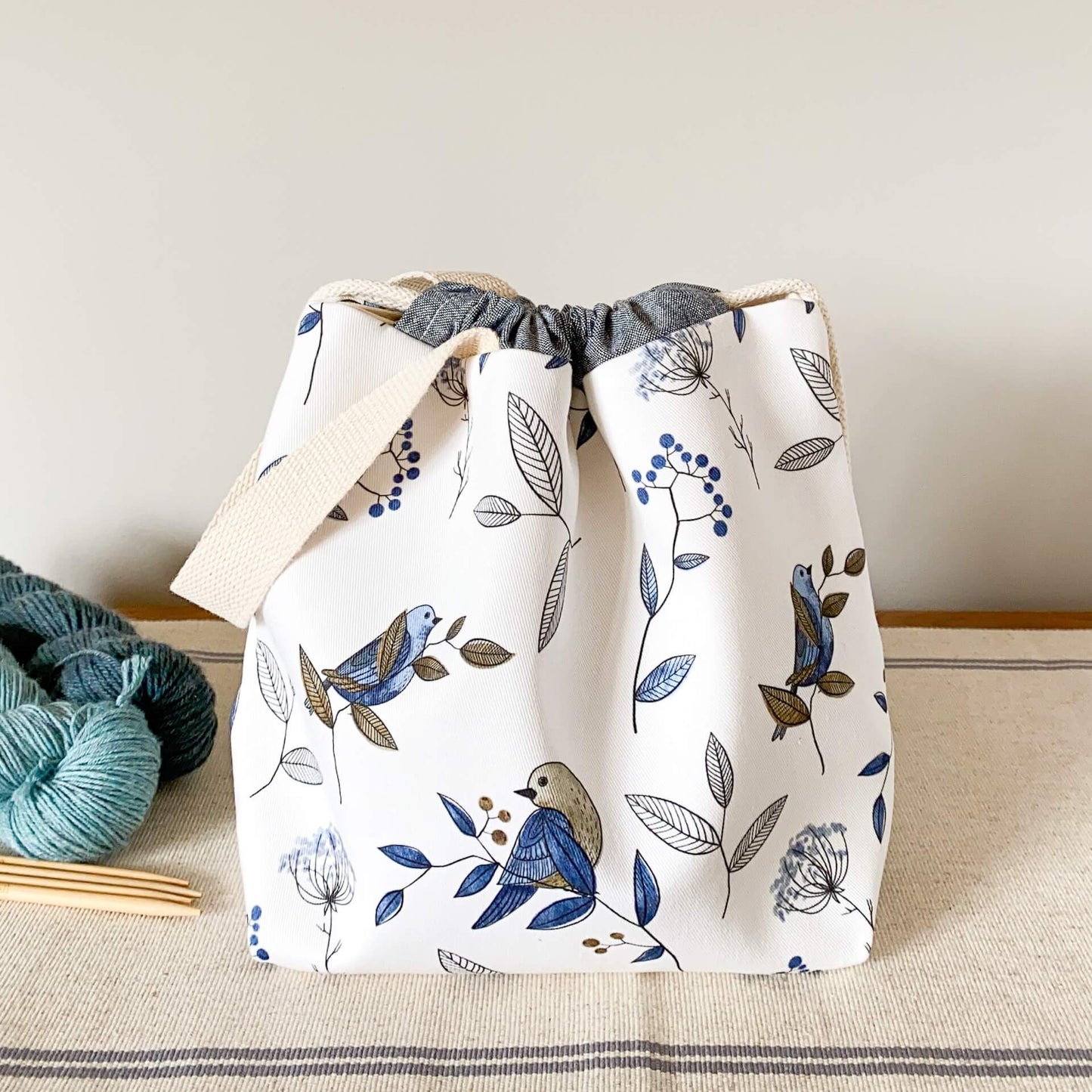 A project bag sits on a table next to skeins of yarn. It is made from a fabric that has been printed with blue birds sitting on branches. 