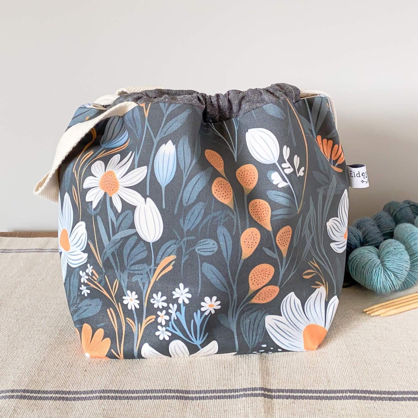 A project bag sits on a table next to two skeins of yarn and wooden knitting needles. The bag is made from a fabric that has a dark background onto whch are printed large scale floral elements. Most of the print is monochrome but there are some orange elements.