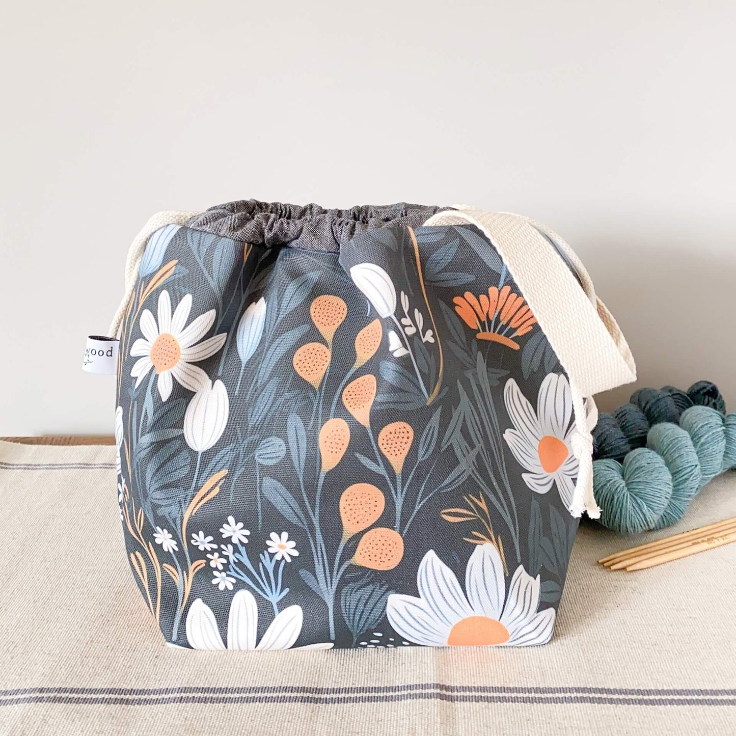 A project bag sits on a table next to two skeins of yarn and wooden knitting needles. The bag is made from a fabric that has a dark background onto whch are printed large scale floral elements.
