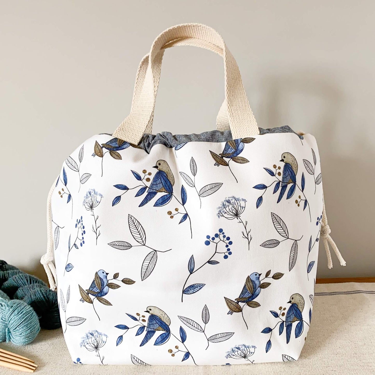 A large knitting project bag, made using a pretty blue bird print, sits on a wooden table next to some yarn and knitting needles. The bag is pulled closed.