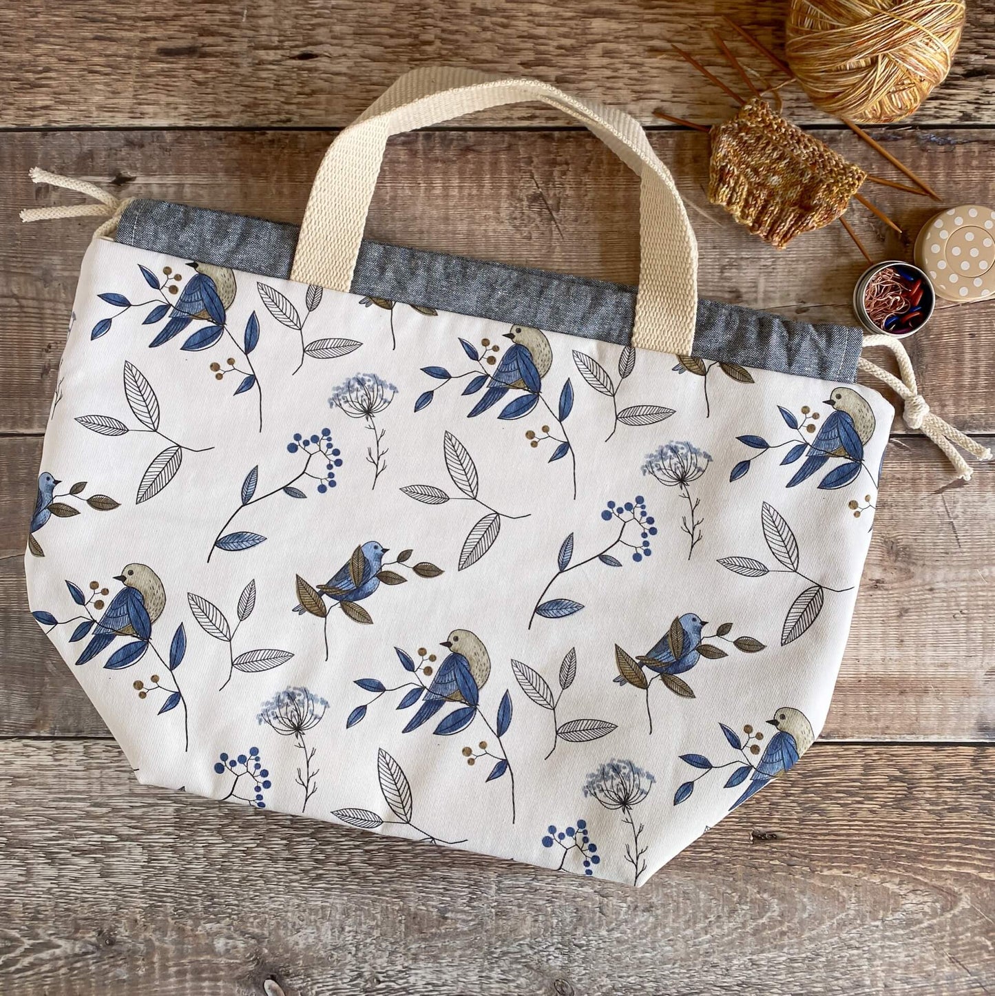 A large knitting project bag with cream handles lies flat on a wooden table next to some in progress knitting. The bag is made with a fabric that is covered in a pretty blue bird print.
