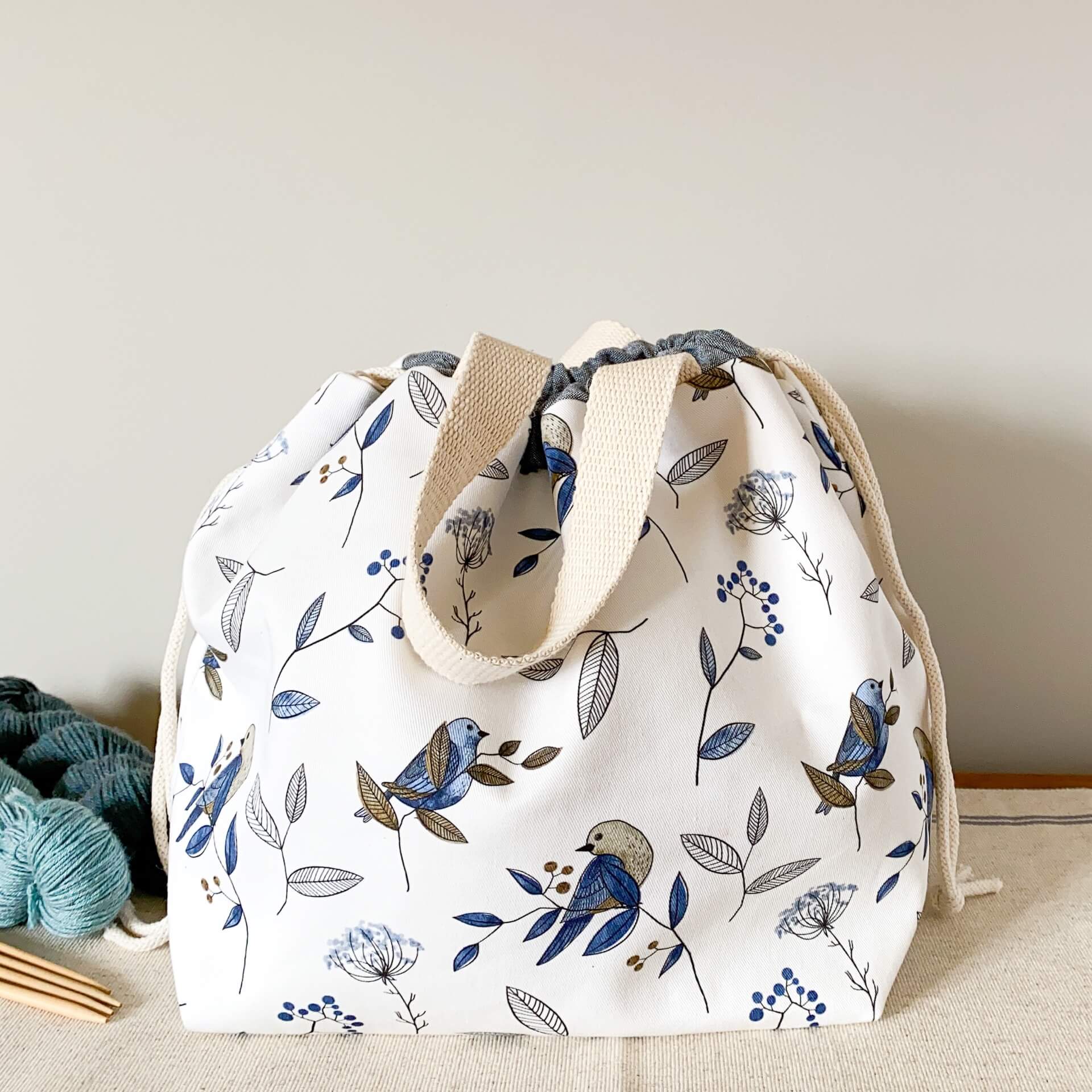 A large knitting project bag, made using a pretty blue bird print, sits on a wooden table next to some yarn and knitting needles. The bag is pulled closed. 
