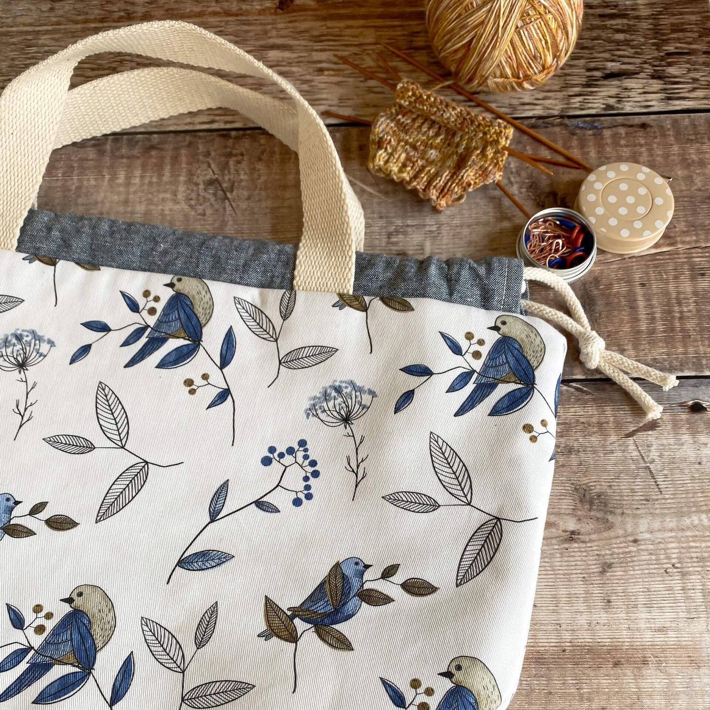 A project bag lies on a wooden table. Just the corner of the bag can be seen alongside a knitting project and some stitch markers.