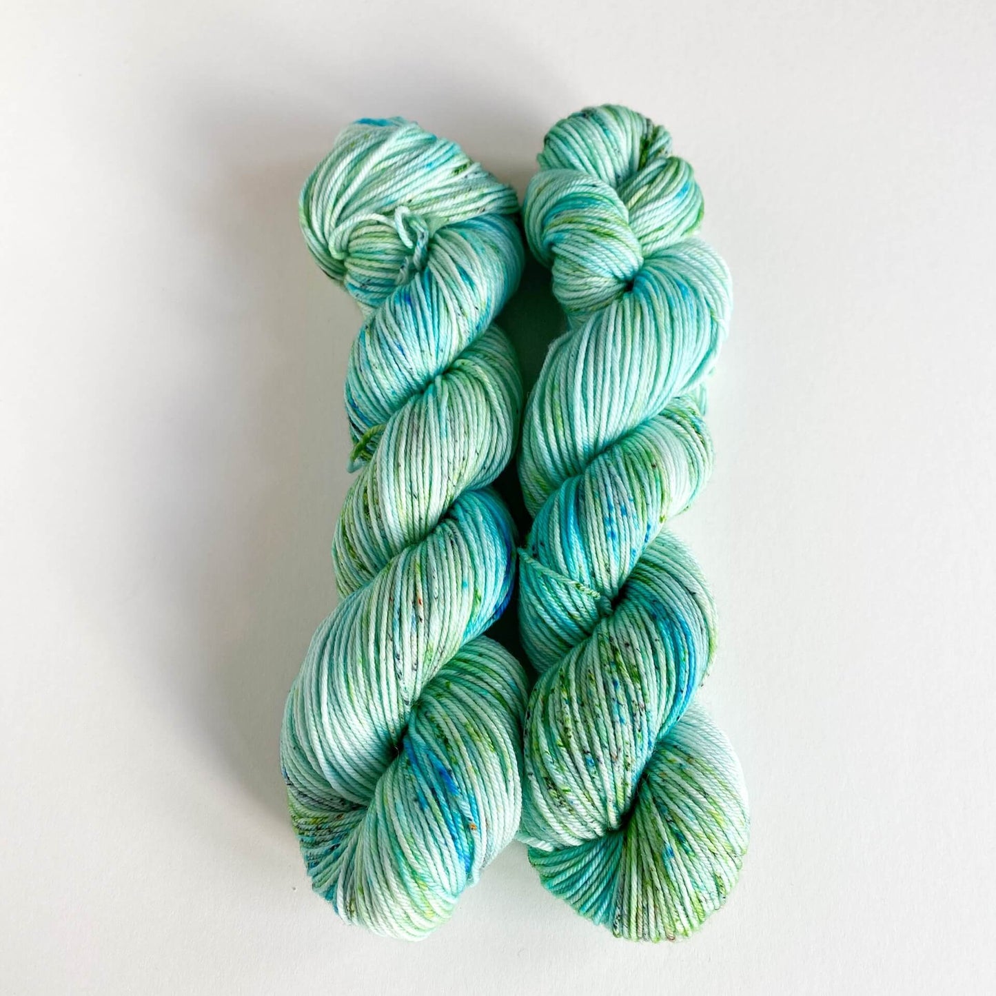 Two skeins of wound yarn on a white background. The yarn is coloured with green and blue dyes and has some speckling.