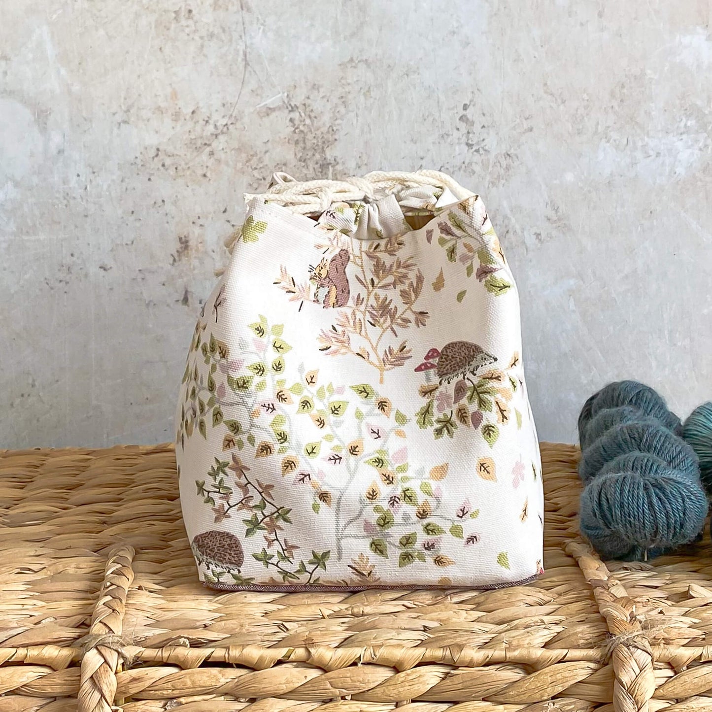 A project bag based on the Japanese rice bag design sits on top of a wicker chest. The fabric is a woodland themed fabric featuring trees and hedgehogs and squirrels.