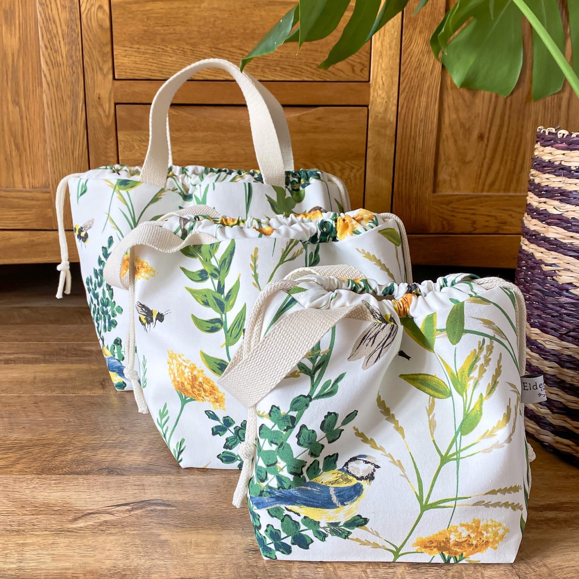 Three knitting project bags sit together with the smallest at the front and the largest at the back. The bags are made from a fabric that shows a summery scene with foliage in bloom, bees and birds. 