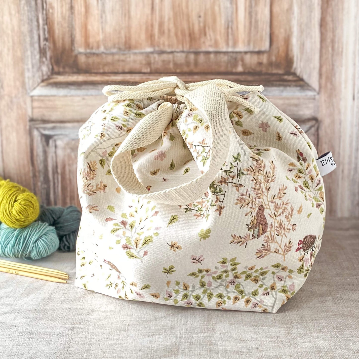 A large knitting project bag with a delicate woodland design featuring birds, hedgehogs, and greenery in soft pastel tones. The bag has fabric handles and a drawstring closure. Nearby, there are skeins of yarn and knitting needles on a neutral surface