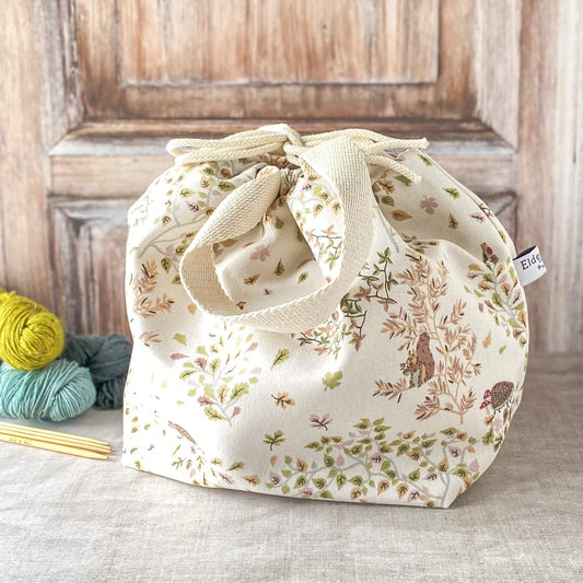 A large knitting project bag with a delicate woodland design featuring birds, hedgehogs, and greenery in soft pastel tones. The bag has fabric handles and a drawstring closure. Nearby, there are skeins of yarn and knitting needles on a neutral surface
