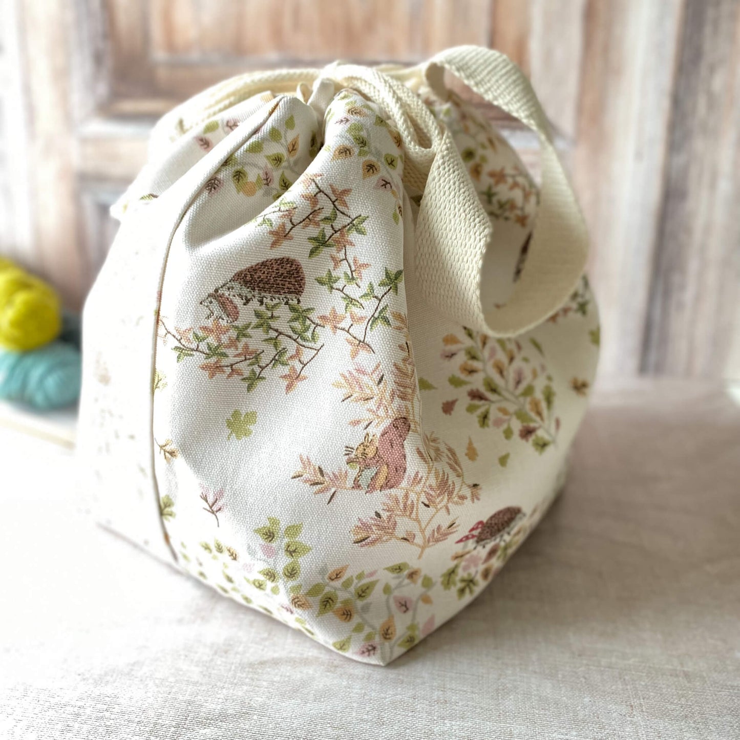 A large knitting project bag featuring a woodland scene with hedgehogs, birds, and leaves in soft pastel colors. The bag has fabric handles and a drawstring closure. Skeins of yarn and knitting needles are visible on the surface next to it.