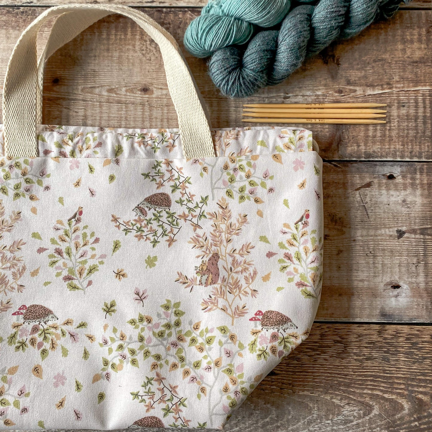 A close-up of a large knitting project bag with a soft woodland design, featuring hedgehogs, squirrels, birds, and leaves in pastel colors. The bag has sturdy fabric handles and a drawstring closure. Nearby are skeins of yarn and knitting needles placed on a wooden surface.