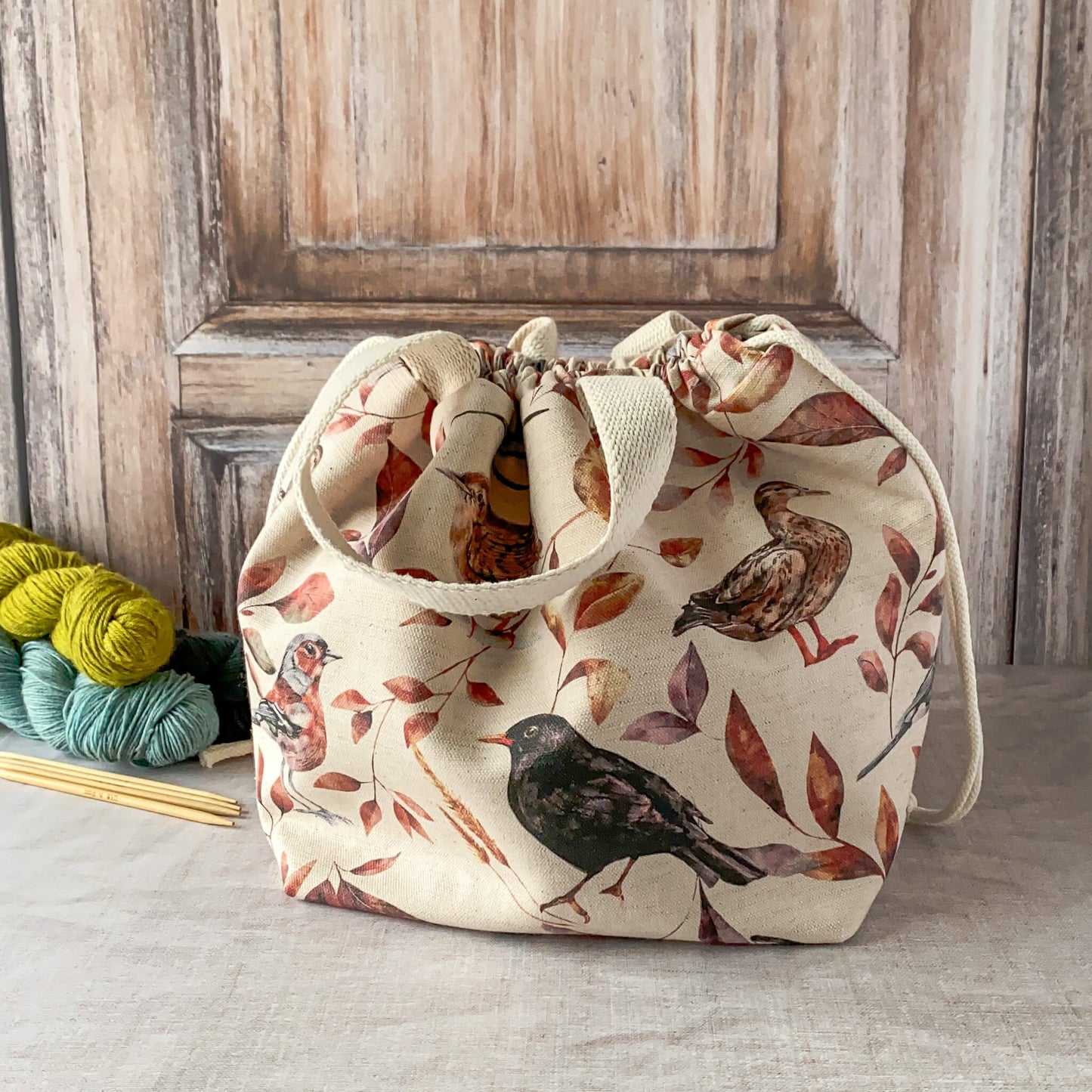This image features a large knitting project bag with a woodland bird and leaf design in earthy tones. The bag is cinched at the top with a drawstring and has sturdy fabric handles. It is placed on a neutral surface with a few skeins of yarn and knitting needles visible nearby. The background has a wooden texture, complementing the natural, rustic aesthetic of the bag.