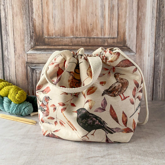This image features a large knitting project bag with a woodland bird and leaf design in earthy tones. The bag is cinched at the top with a drawstring and has sturdy fabric handles. It is placed on a neutral surface with a few skeins of yarn and knitting needles visible nearby. The background has a wooden texture, complementing the natural, rustic aesthetic of the bag.