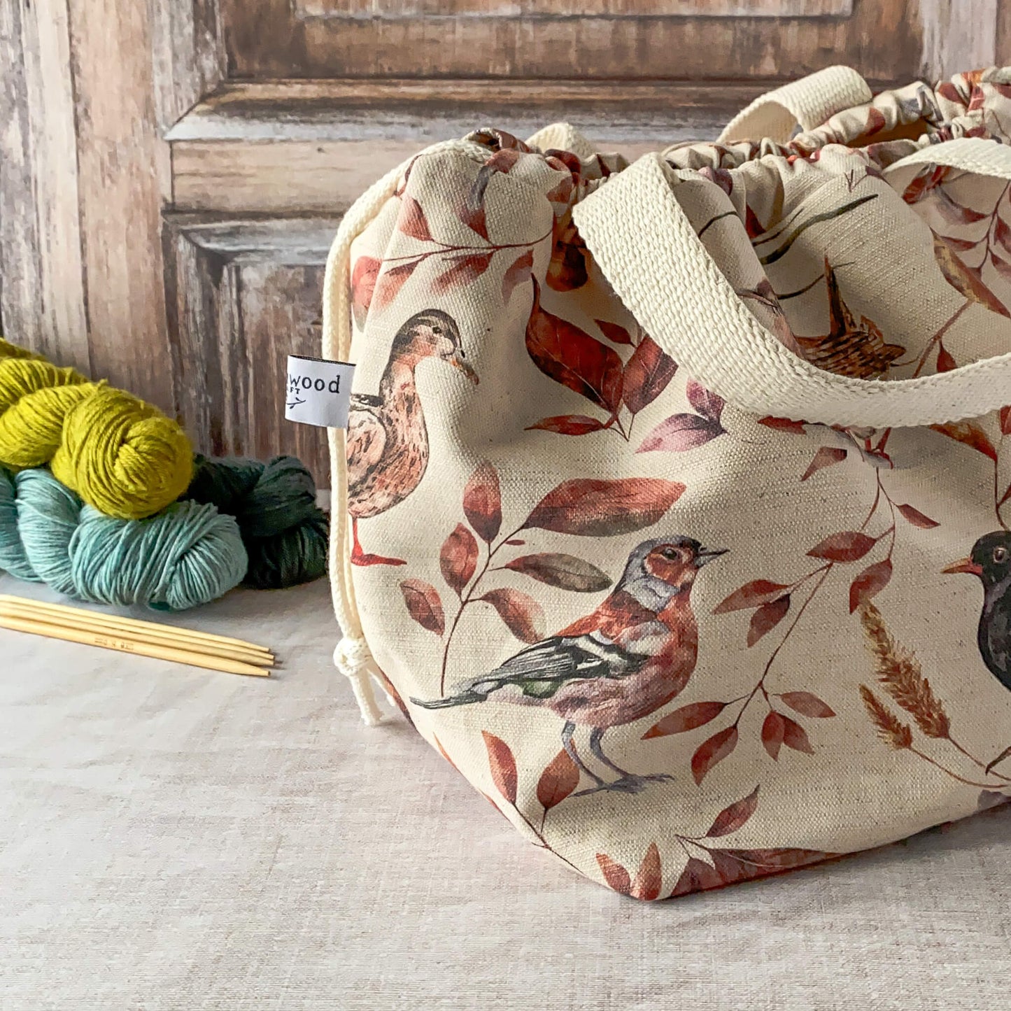 This image shows a large knitting project bag with a woodland bird and leaf print in earthy tones. The bag is cinched with a drawstring and has fabric handles. Nearby are skeins of yarn and knitting needles on a neutral surface.