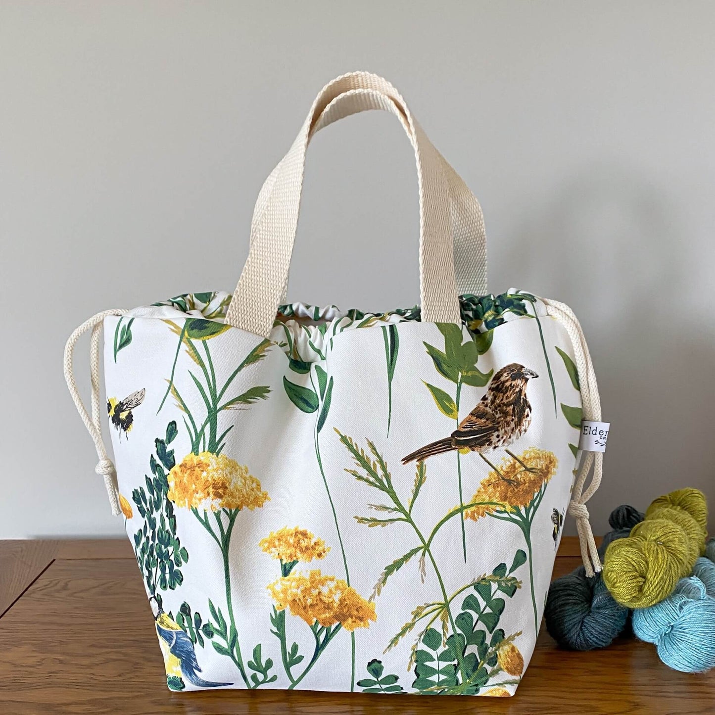A large drawstring bag sits on a wooden table with its handles pulled up. The bag is next to three skeins of yarn. Printed on the bag is a summery nature scene with orange foliage, birds and bees.