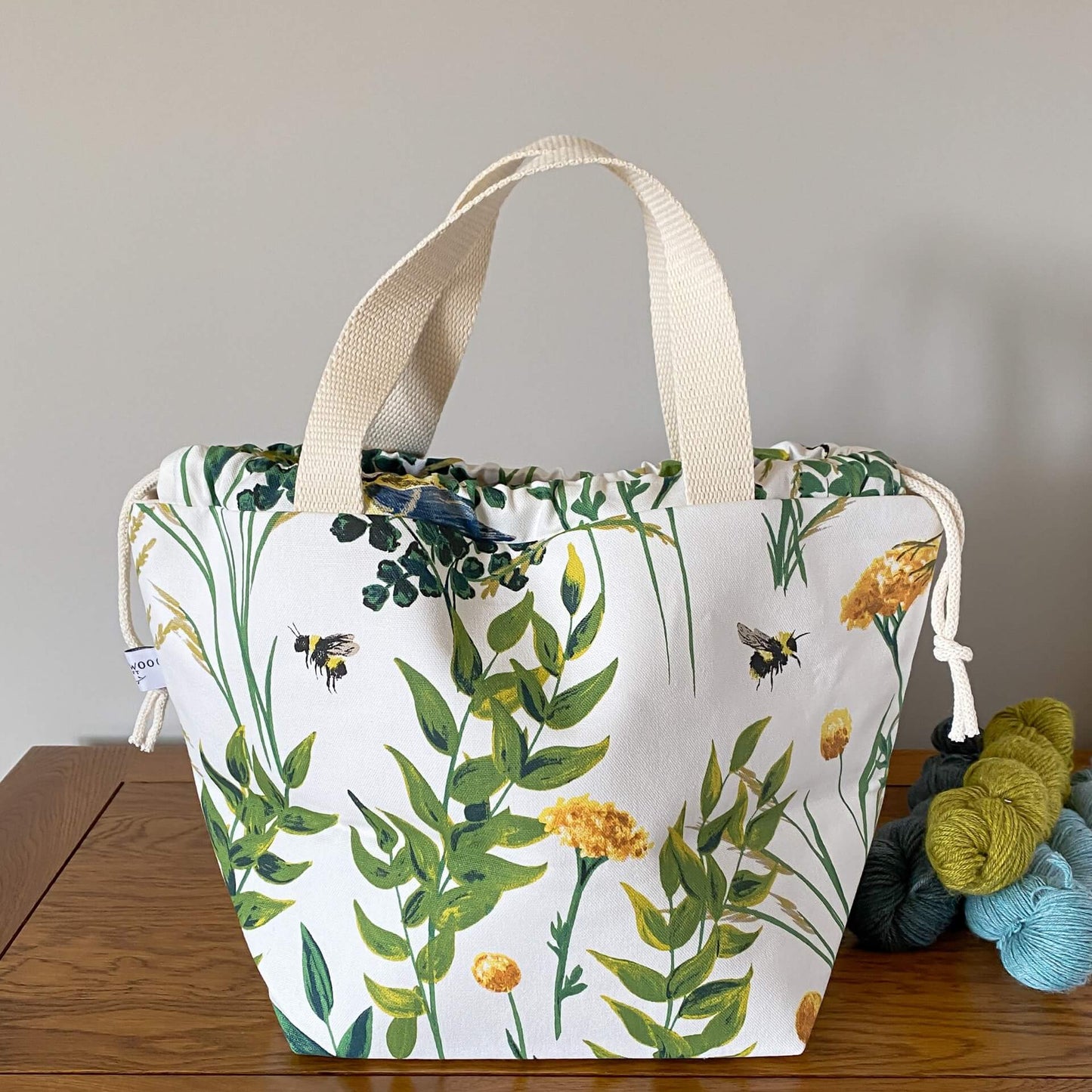 A large drawstring bag sits on a wooden table with its handles pulled up. The bag is next to three skeins of yarn. Printed on the bag is a summery nature scene with orange foliage and bees.