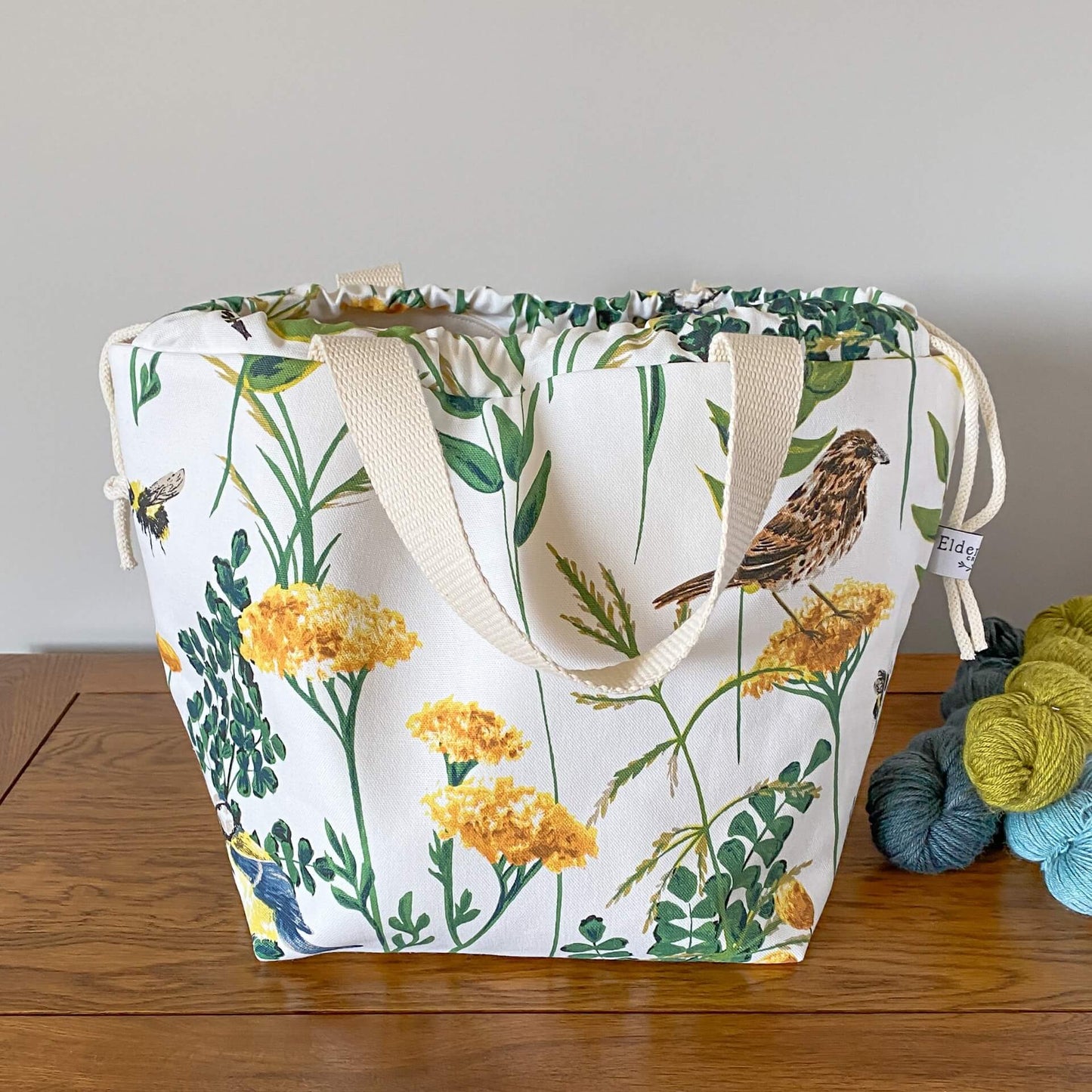 A draswtring bag is seen on a table next to some yarn. The handles of the bag hang down. The bag is made from a print of a summery scene with foliage and birds and bees. On this bag there is a song thrush and a blue tit. 