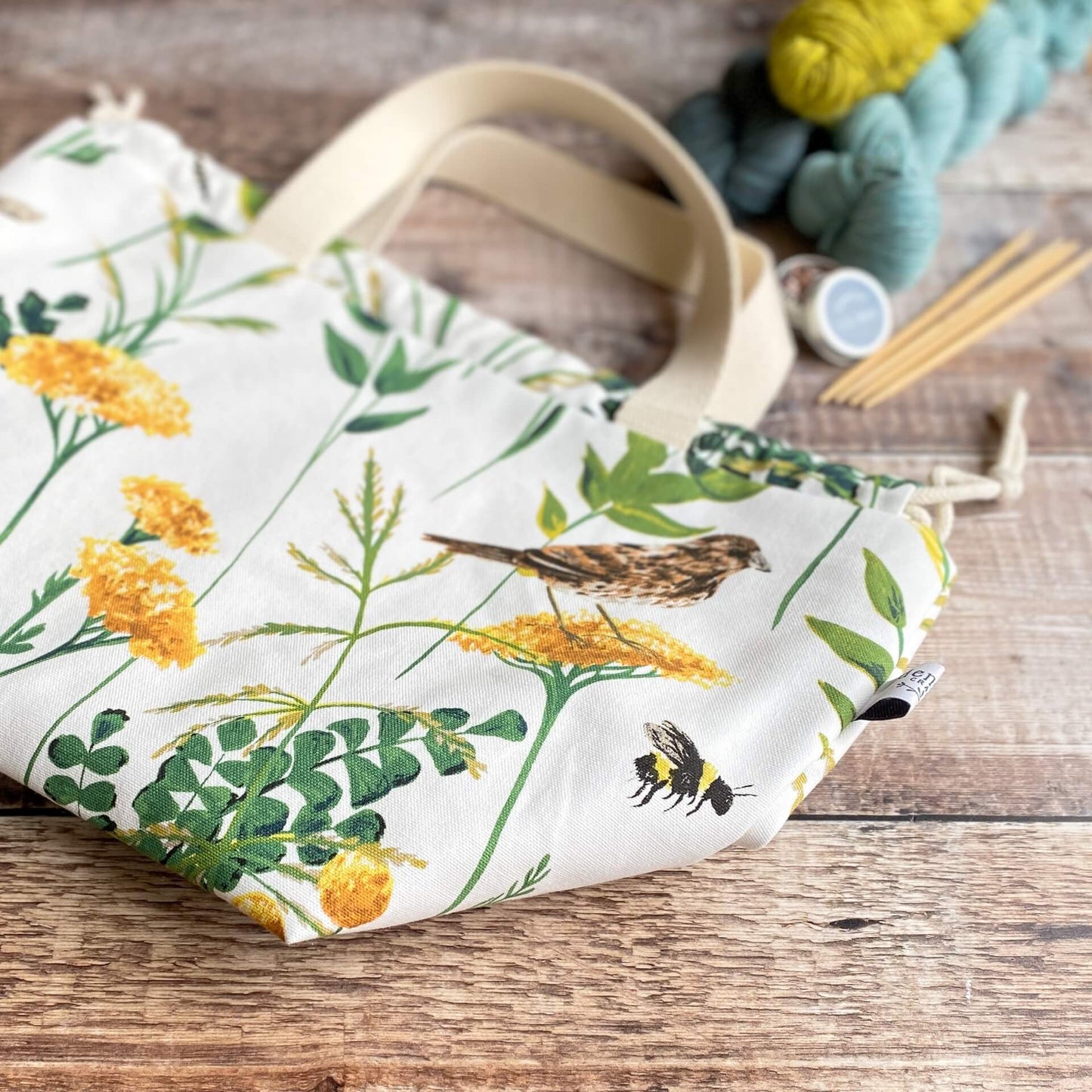 A project bag lies flat on a wooden table. Next to the bag is some yarn, four wooden knitting needles and an open tin containing stitch markers.